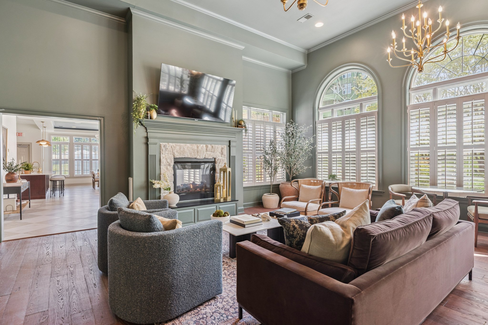 Living room with large arched windows, a fireplace with a TV above, multiple chairs and a sofa, and a chandelier.