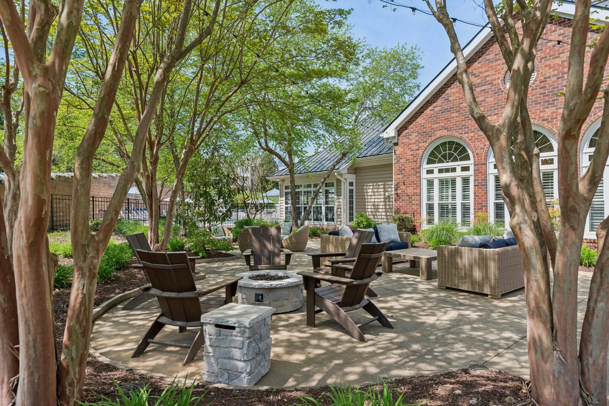 A backyard patio with outdoor seating around a fire pit, surrounded by trees and a brick house with large windows.