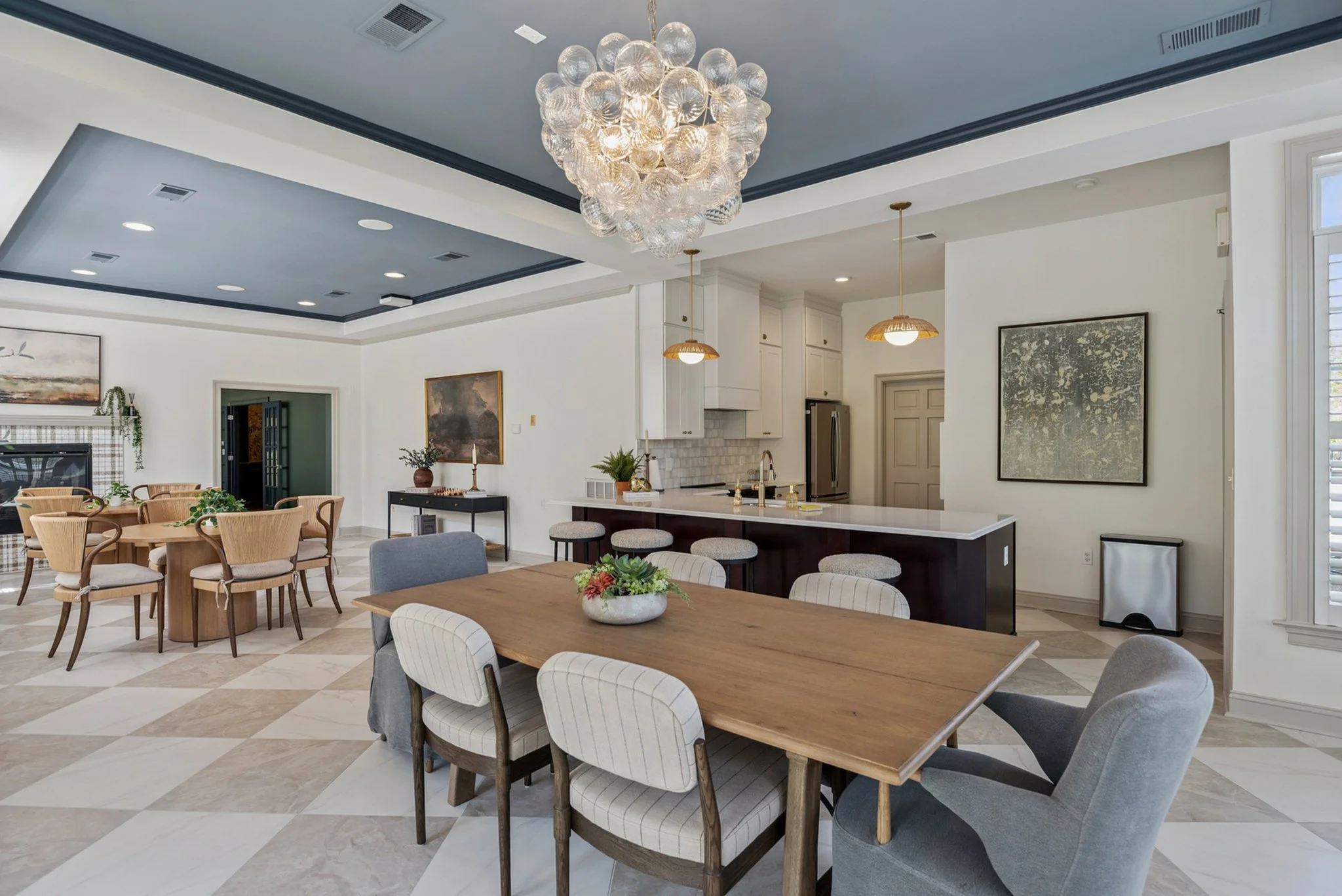 Interior view of a modern kitchen and dining area with a large chandelier, light-colored cabinets, a dark island with barstools, and around a central wooden dining table with chairs around it.