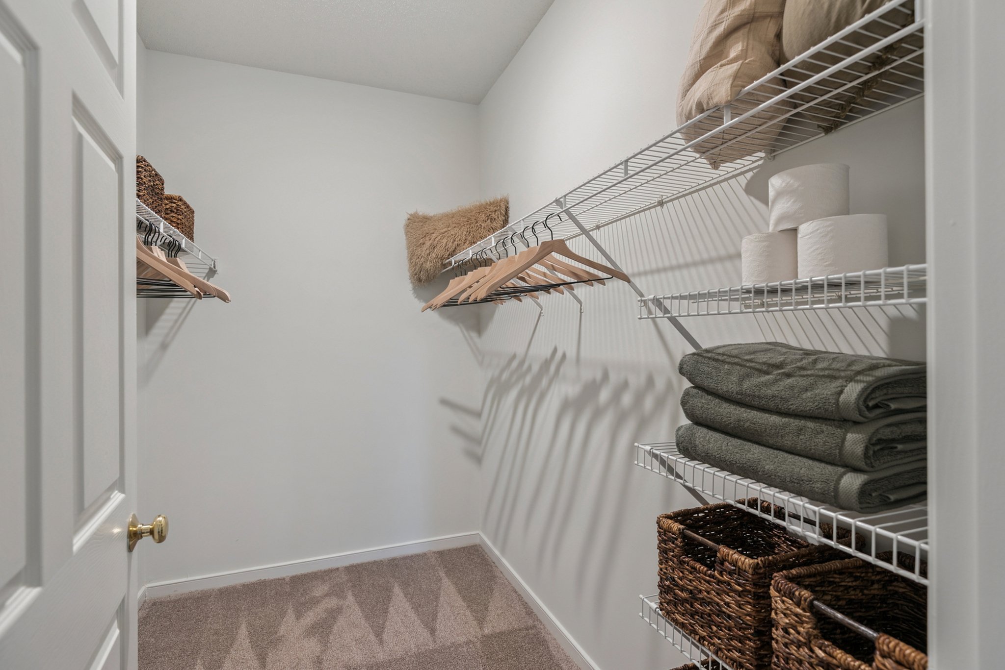 Empty closet with white wire shelves holding folded towels, baskets, toilet paper rolls, and clothing hangers on a beige carpeted floor.