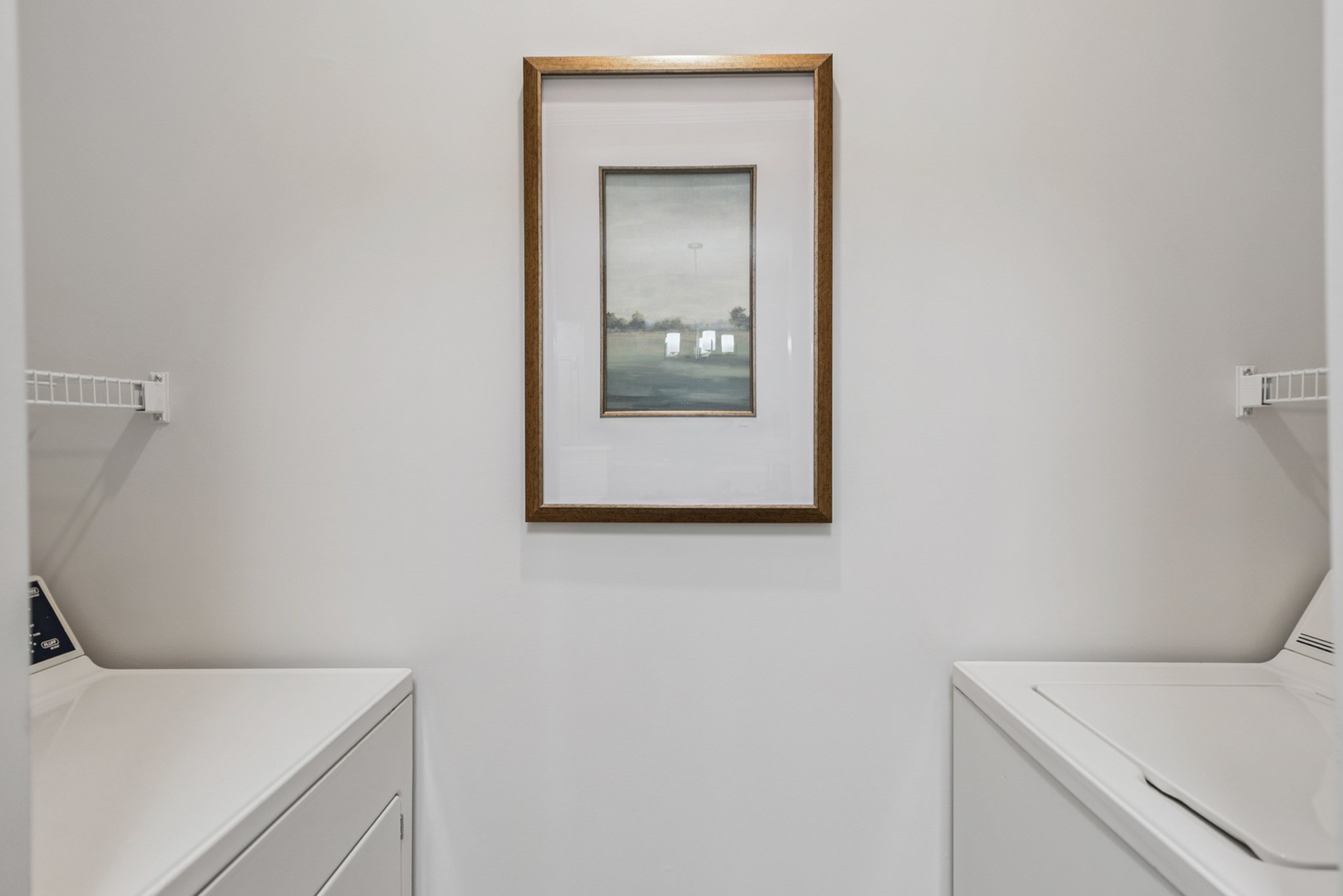 A laundry room with white walls, a framed landscape painting, and two white washing machines on either side.