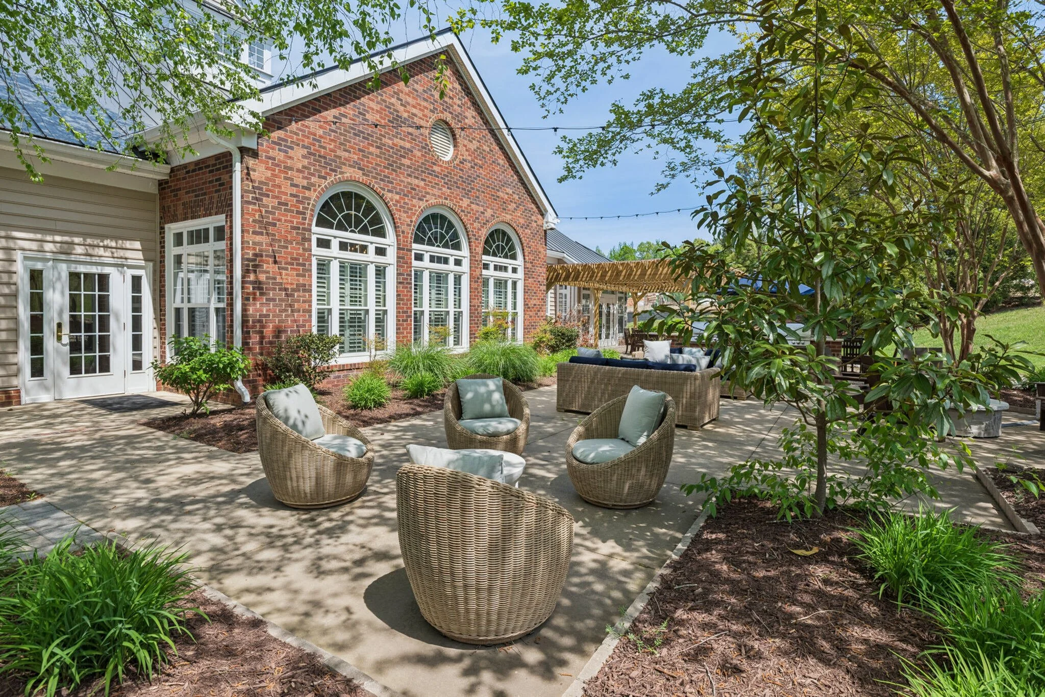 A patio area with wicker chairs and a sofa on a concrete surface, surrounded by trees and plants, in front of a brick house with large arched windows and a string of outdoor lights.