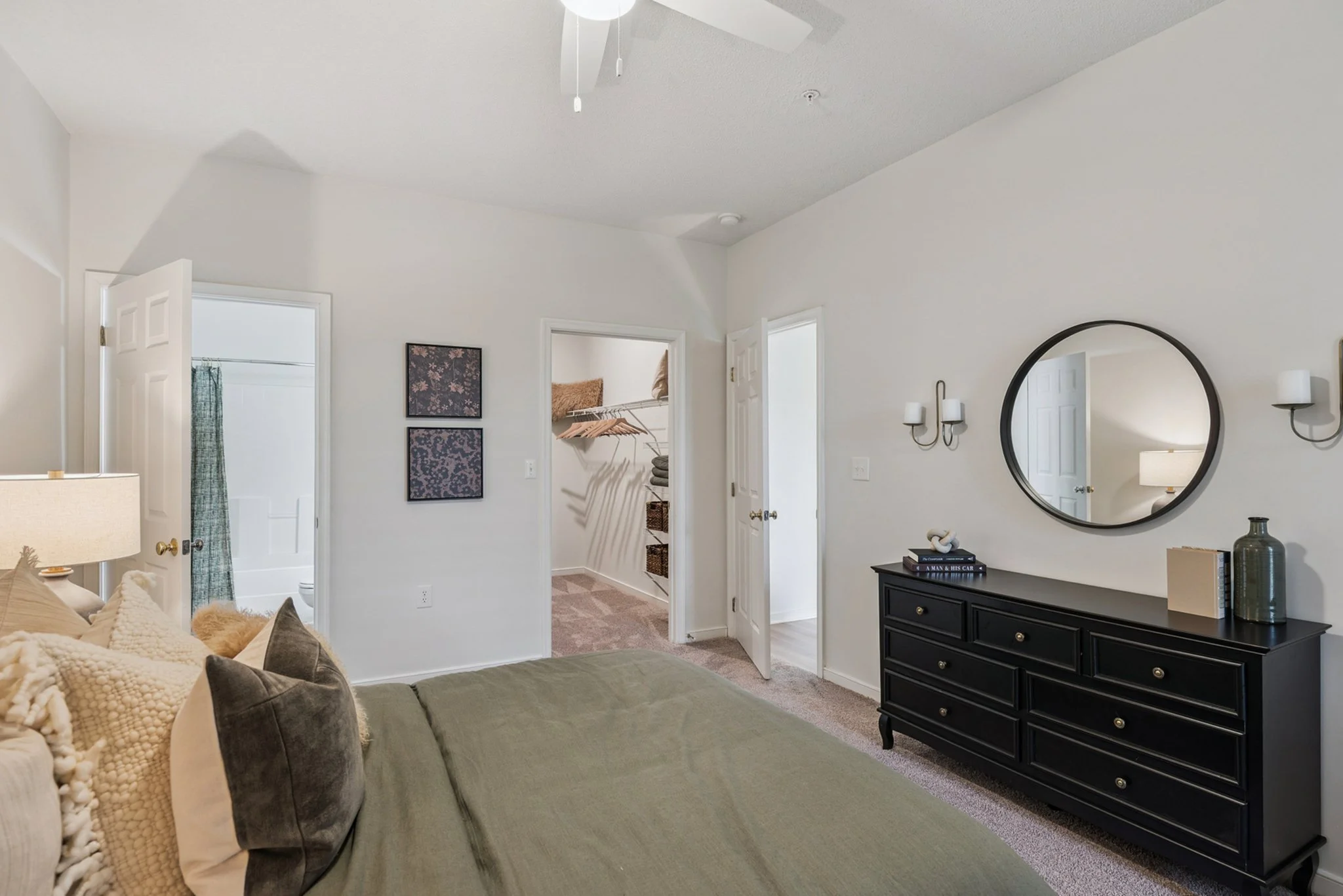 Living room with a gray-green sofa, various pillows, a black dresser with a mirror above, and two wall-mounted candle holders. An open door reveals a closet with shelves and hanging clothes. In the background, an open door leads to a bathroom with a 