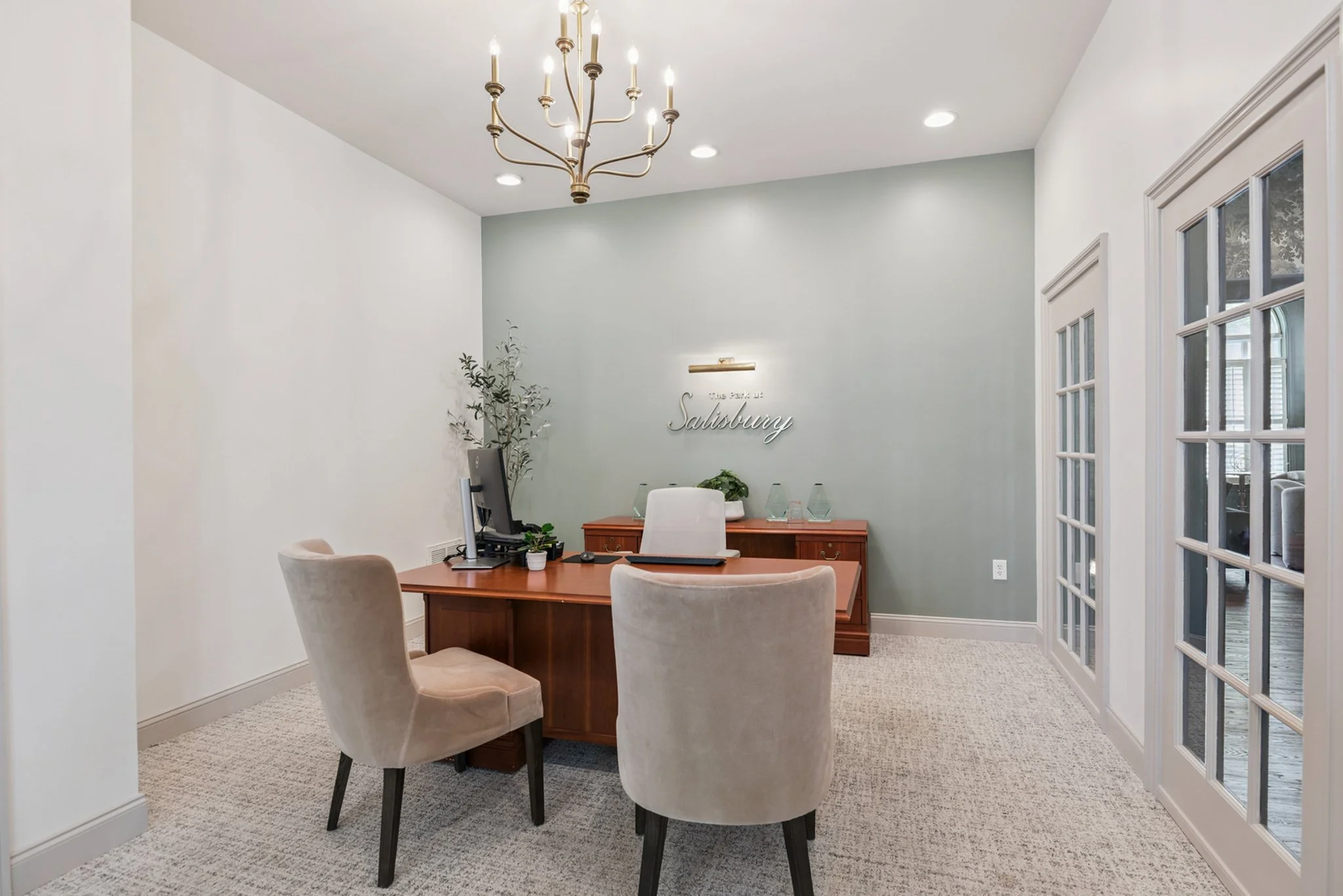 An office with a wooden desk, three beige upholstered chairs, a computer, potted plants, a wall sign reading 'The Park at Salisbury,' a modern chandelier, and a glass-paneled door.