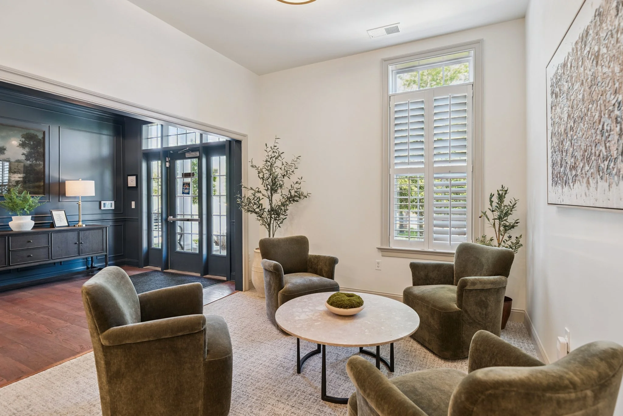 Living room with high ceiling, large windows with shutters, and seating area with multiple armchairs surrounding a round coffee table with a decorative bowl. There is a side table with a lamp and a piece of artwork on the wall.