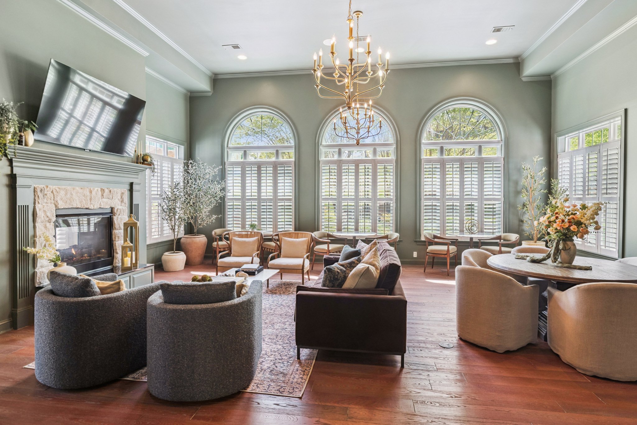 Living room with large arched windows with white shutters, a chandelier, a fireplace with a TV mounted above, and various seating including sofas and armchairs, decorated with plants and flowers.