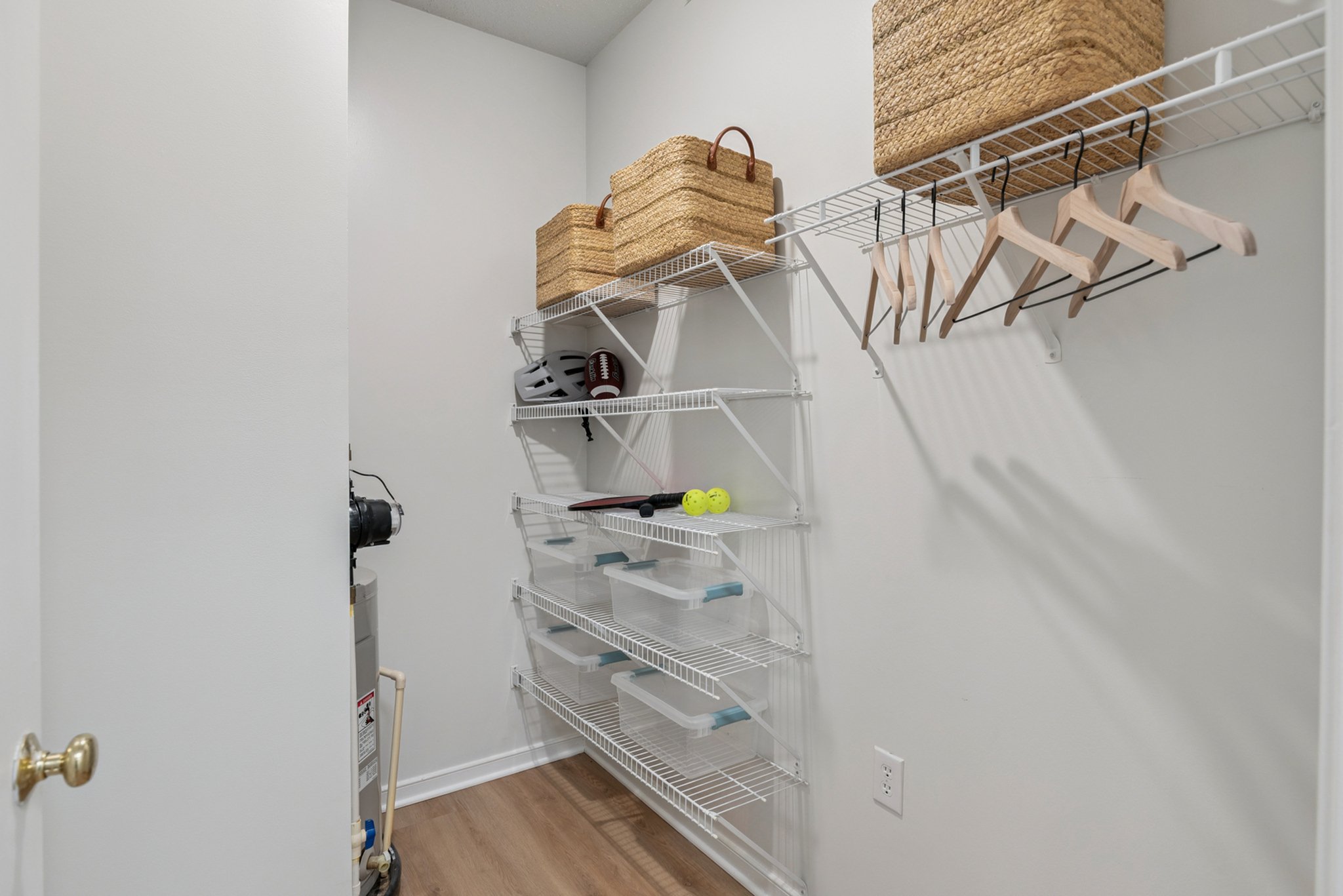 Empty white wire shelves on a wall, with woven baskets, wooden hangers, tennis balls, and a football. The room has a brown hardwood floor and white walls.