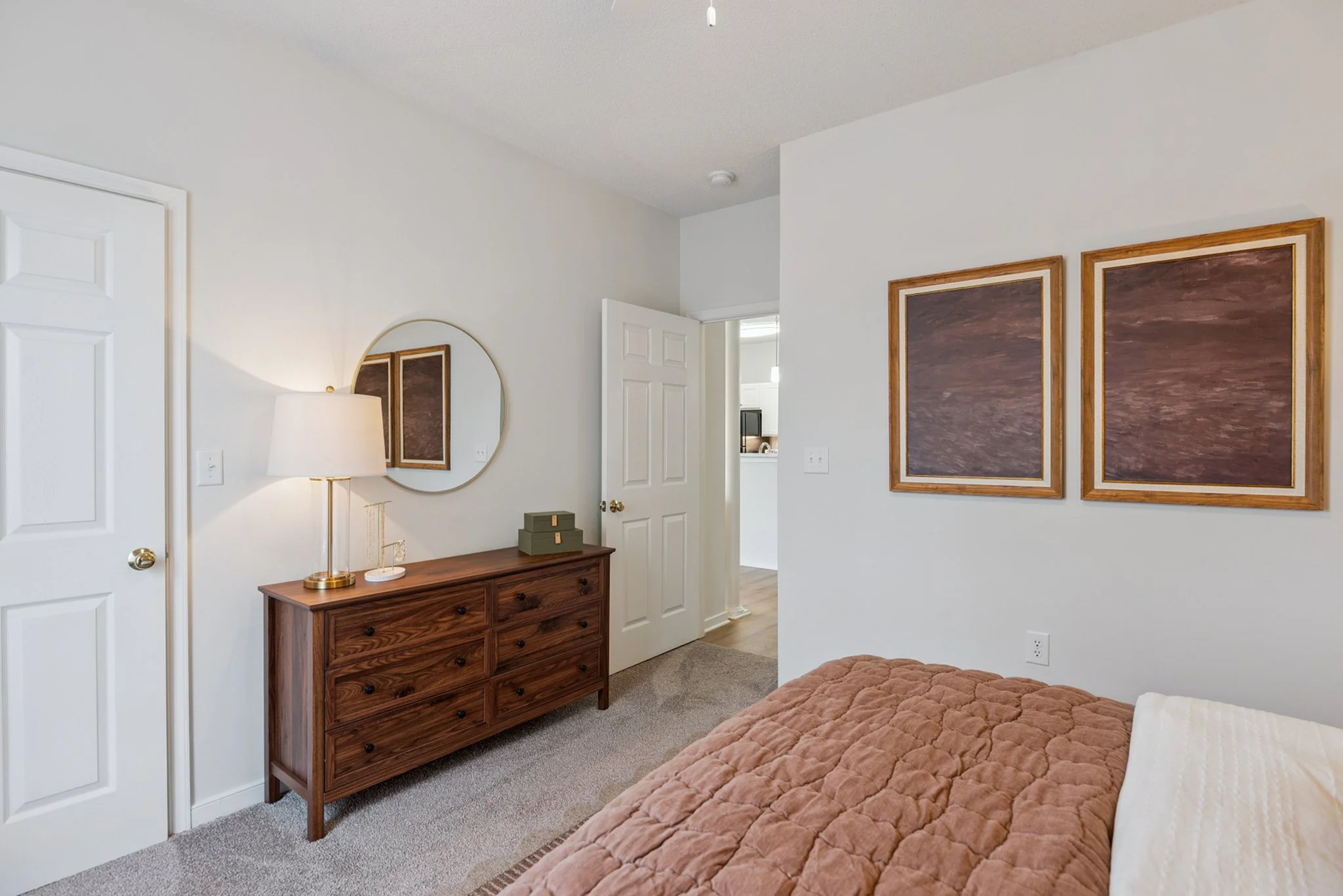 A cozy bedroom with a bed covered in a brown quilt, a wooden dresser with a mirror on the wall, and framed artwork. There is a doorway leading to another room and a bedside lamp on the dresser.
