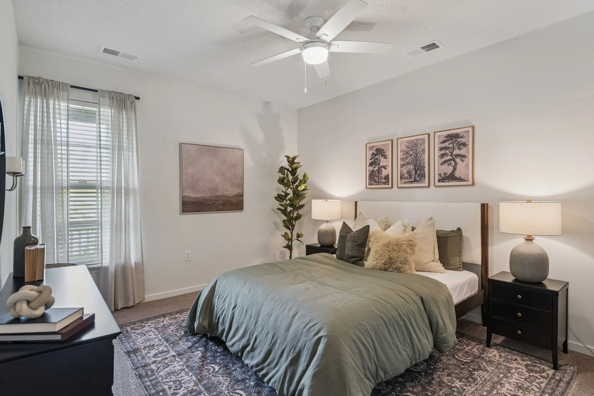 A cozy bedroom with white walls and a ceiling fan, featuring a bed with green bedding, beige and gray pillows, and matching nightstands with lamps. There are framed nature prints above the bed, a large window with sheer curtains, and decorative objec