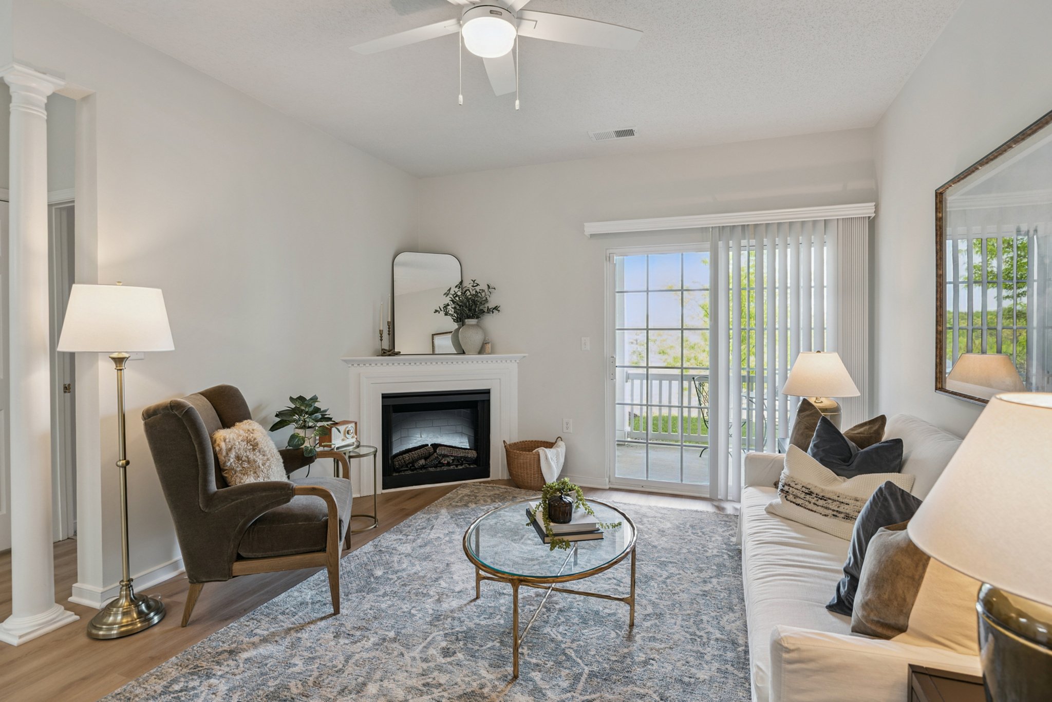 Living room with white walls, a white fireplace with a mirror and decorative items above it, a sliding glass door with vertical blinds, a ceiling fan, a sofa with pillows, two armchairs, side tables with lamps, a glass coffee table with books and a p
