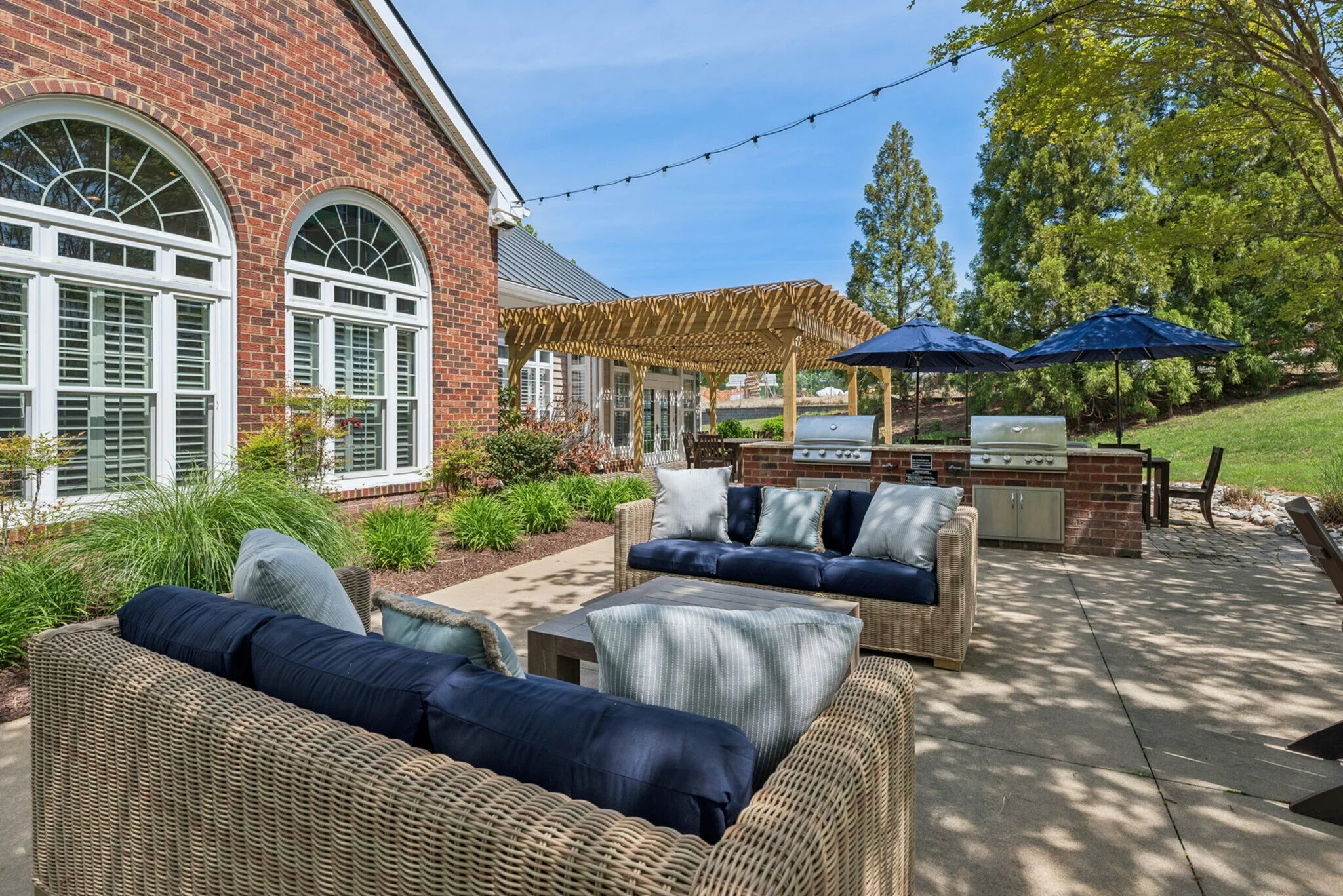 A backyard patio with wicker furniture, blue cushions, and pillows, shaded by trees and string lights, in front of a red brick house with arched windows and a pergola.