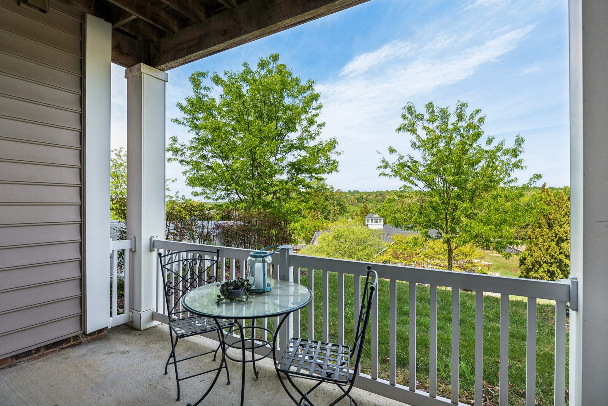 Outdoor balcony with a round glass table, three metal chairs, and decorative items, overlooking green trees and a cloudy sky.