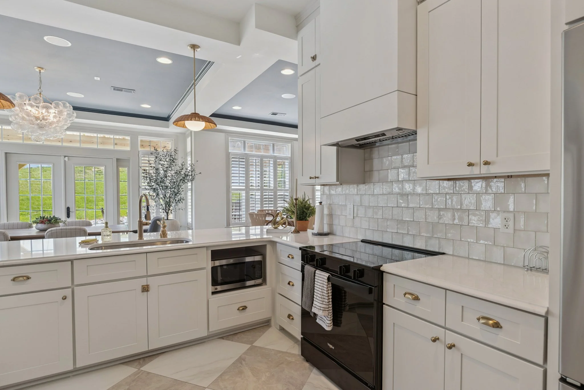 Modern white kitchen with black appliances, gold hardware, subway tile backsplash, large windows with shutters, and view of a dining area with chairs and greenery outside.