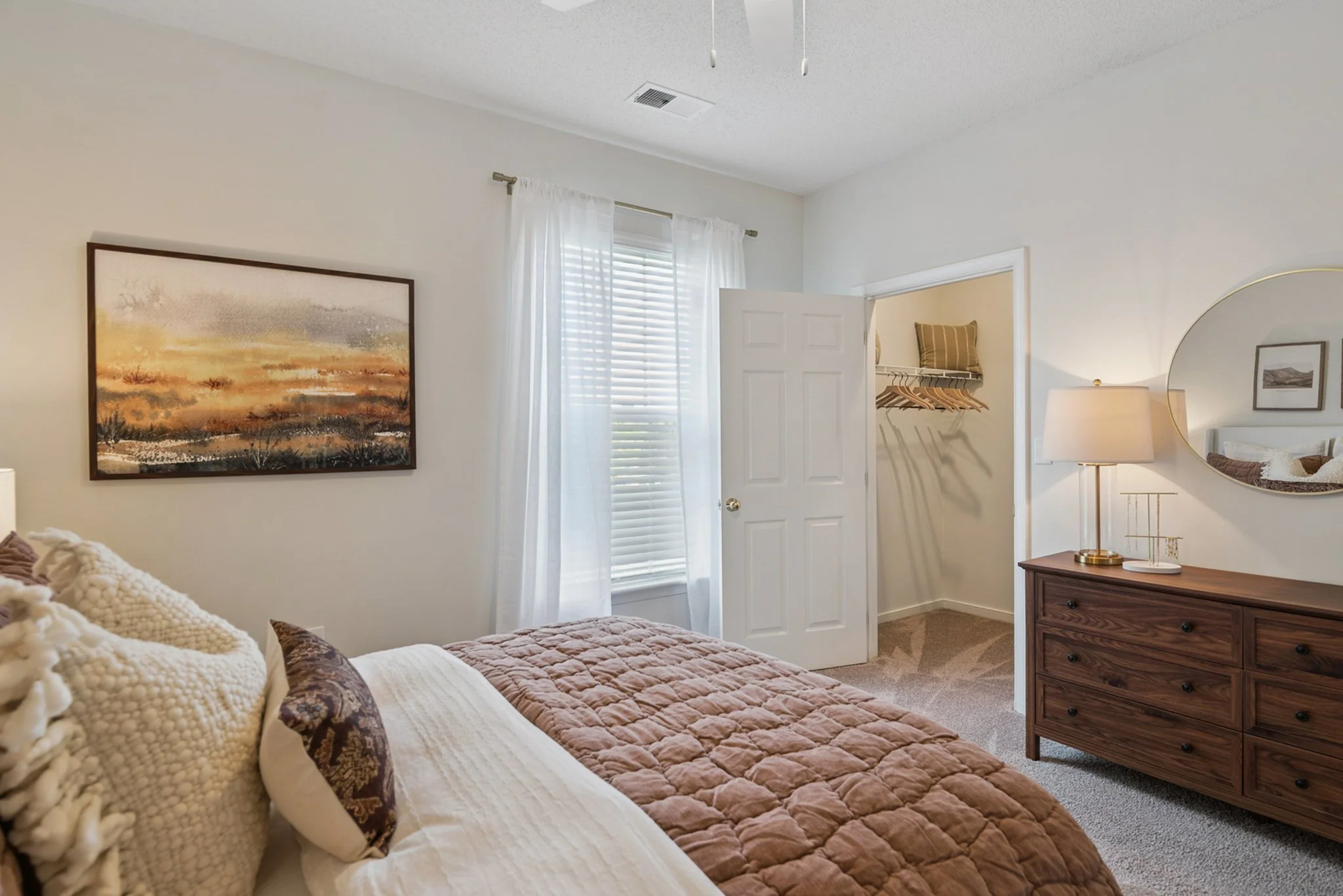 A bedroom with a bed, decorative pillows, a framed landscape picture, a window with white curtains, a dresser, a wall mirror, and a small open closet with hangers and a pillow.