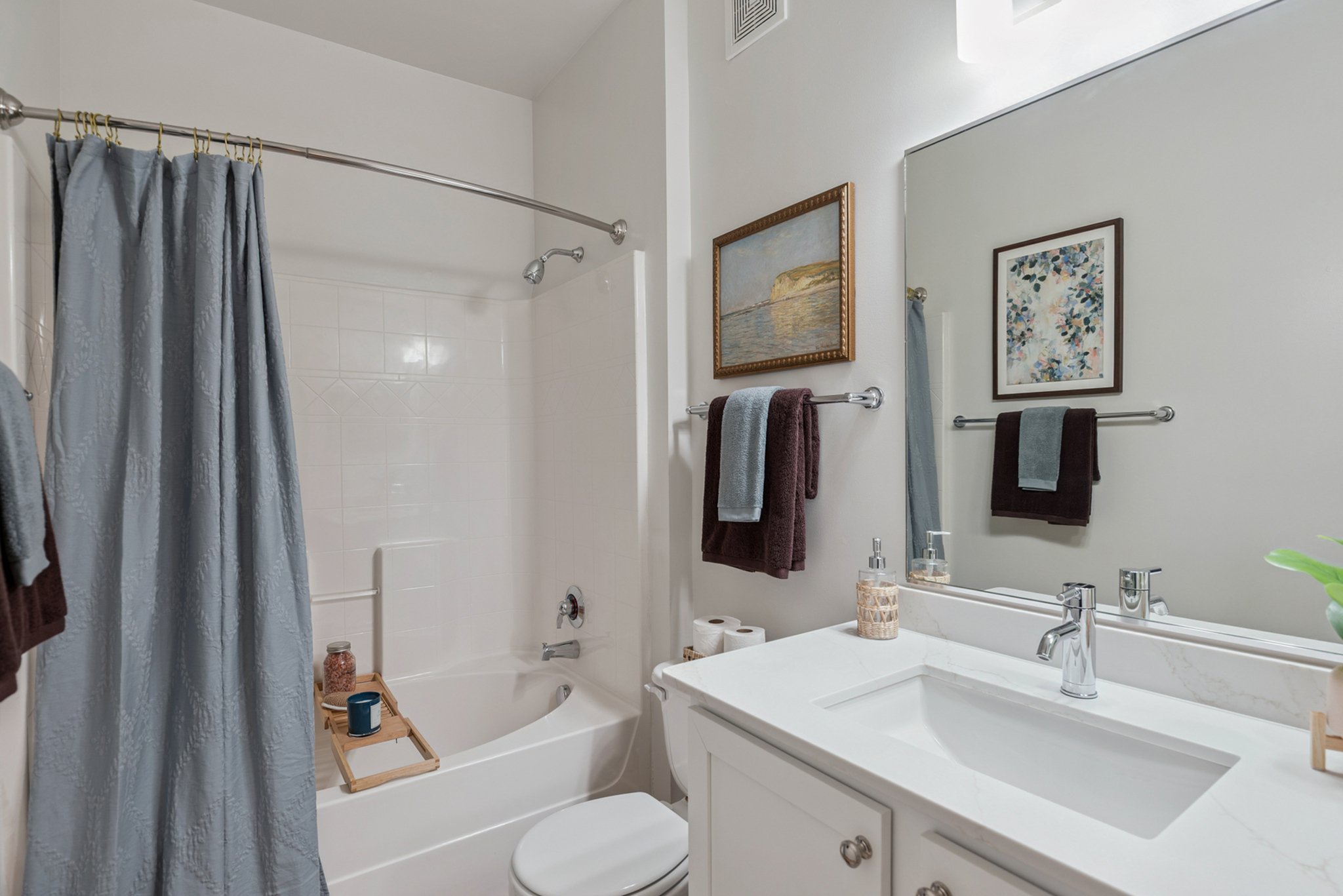 A clean bathroom with a bathtub partially enclosed by a light blue shower curtain, white tiled walls, and a white vanity with a large mirror. Decor includes framed artwork and neatly arranged towels.
