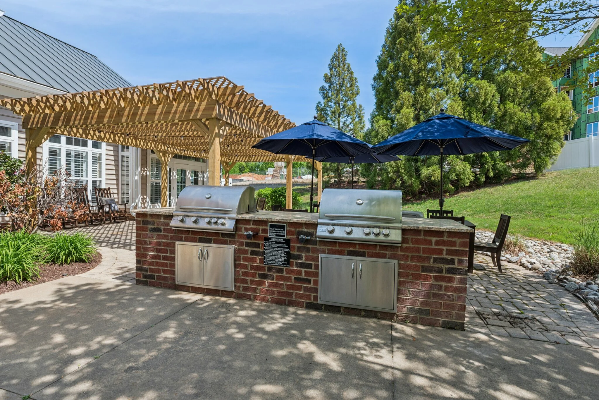 An outdoor area with a brick barbecue grill station, two closed blue umbrellas, and a wooden pergola with lattice design. There are chairs, plants, trees, and a grassy yard in the background.