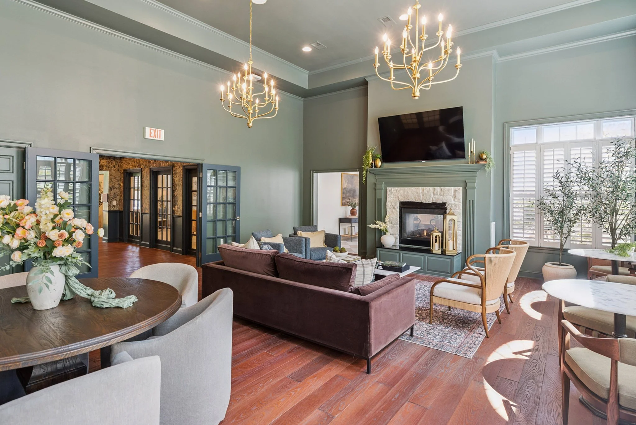 Bright living room with high ceilings, two gold chandeliers, a stone fireplace, a large television, various seating including a sofa and chairs, large windows with white shutters, and decorative plants.