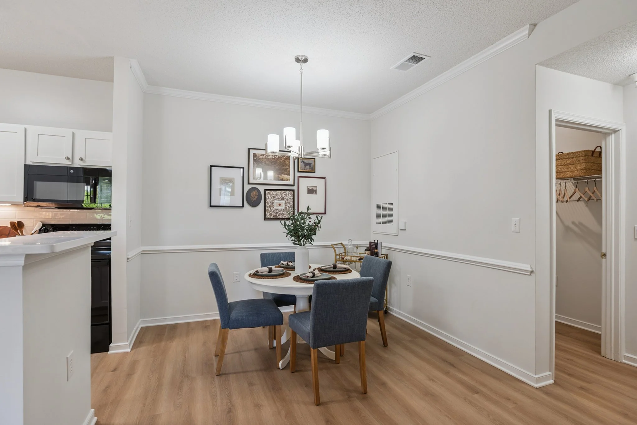 Dining area with a round table set for four, surrounded by four blue upholstered chairs, a pendant light hanging above, white walls with framed artwork, and a small closet in the background.