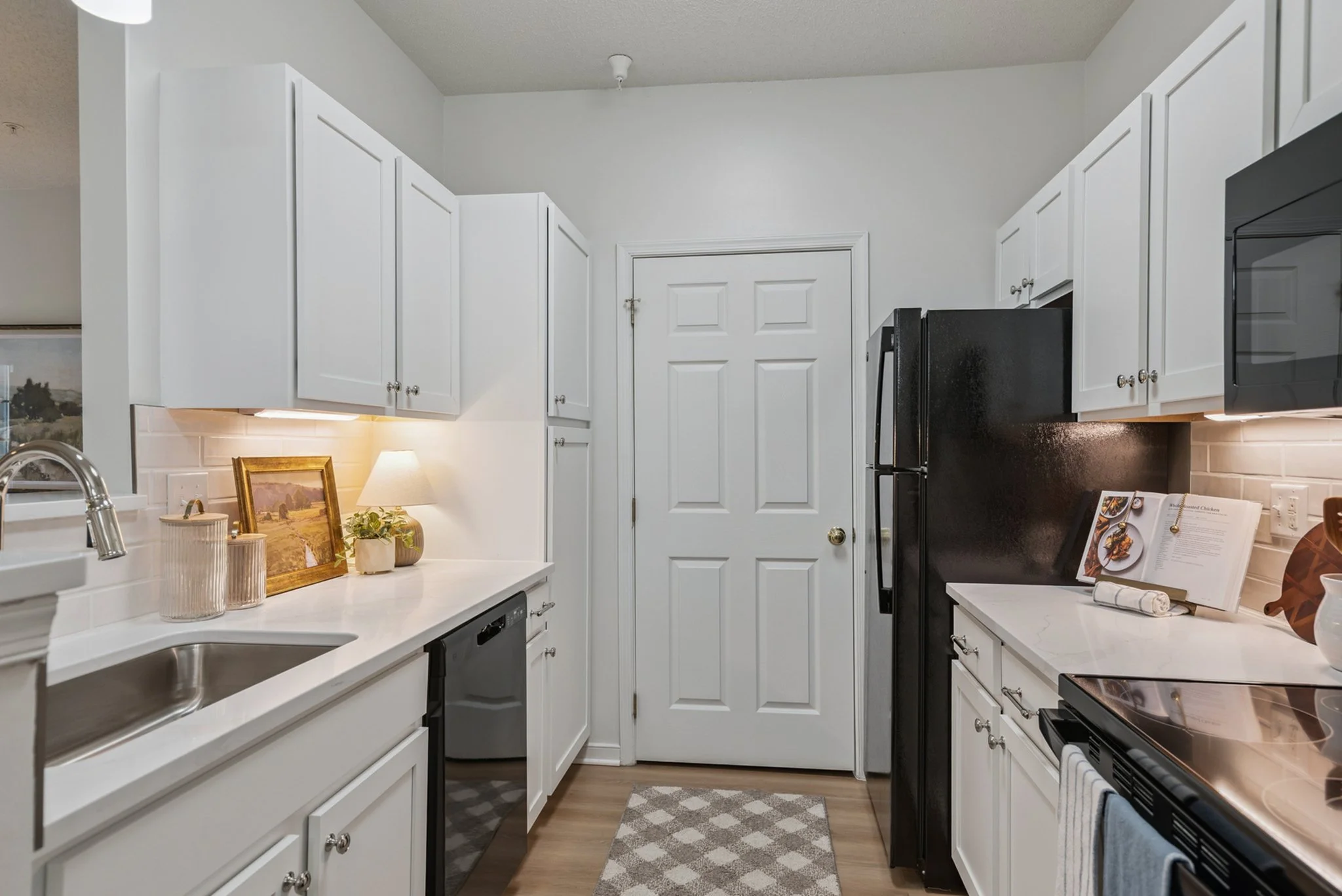 A kitchen with white cabinets, black appliances, a white countertop, and framed artwork. There are decorative items, a lamp, and a recipe book on the counter.
