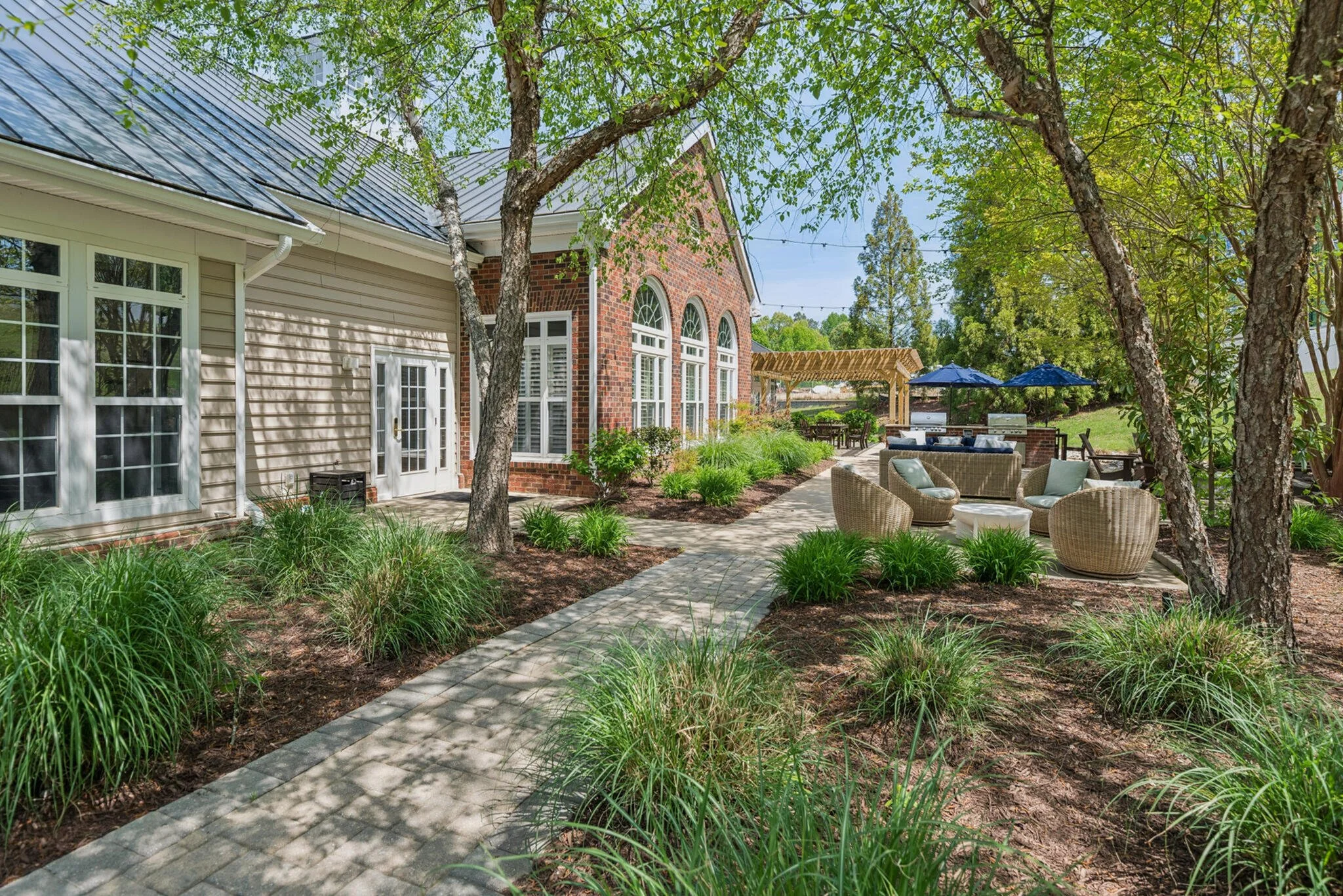 A backyard patio with outdoor seating, including a wicker sofa and chairs, shaded by trees, with umbrellas and string lights, adjacent to a brick and beige house with large windows.