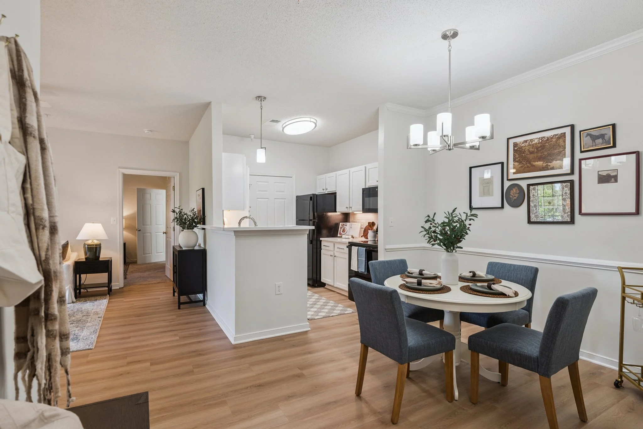 Open-concept living area and kitchen with white walls, mounted pictures, gray dining chairs, and wood flooring.