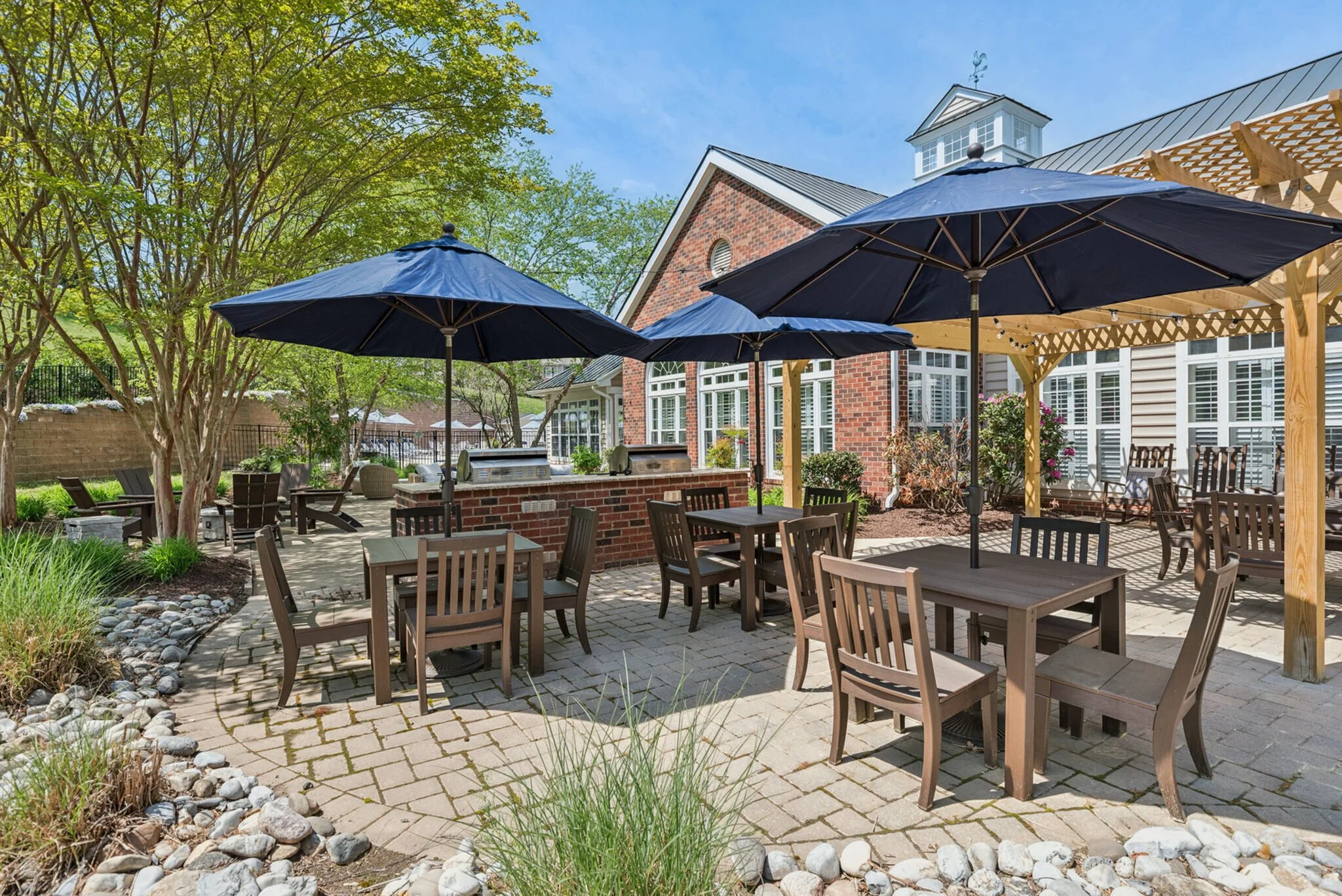 Outdoor patio area with wooden tables and chairs, large navy umbrellas, a brick wall with built-in grills, and greenery with trees in the background.