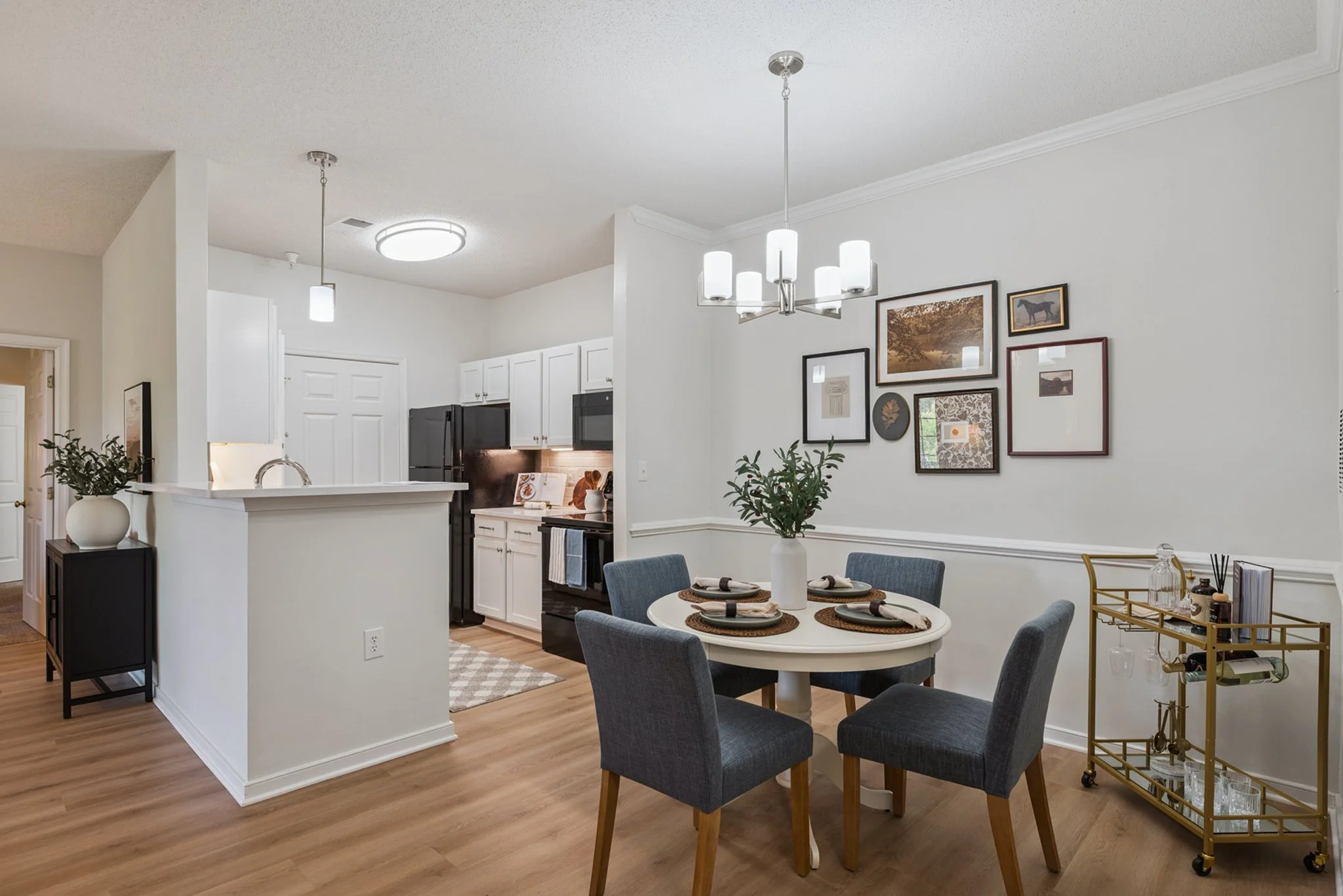Open-concept kitchen and dining area with a white wall, modern chandelier, gallery wall artwork, and a round dining table with four blue chairs, featuring a vase with green foliage.