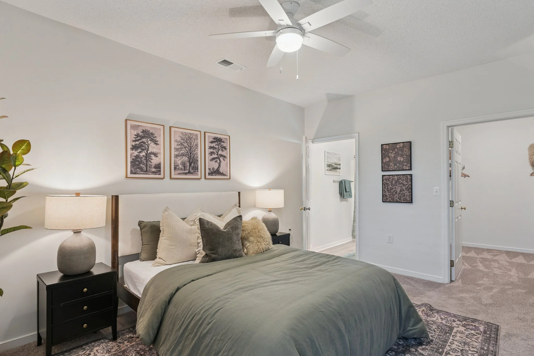 A cozy bedroom with a bed covered in a dark green bedspread, surrounded by pillows. On each side of the bed are black nightstands with large ceramic lamps. Decor on the wall includes three black and white framed pictures of trees. To the right, there
