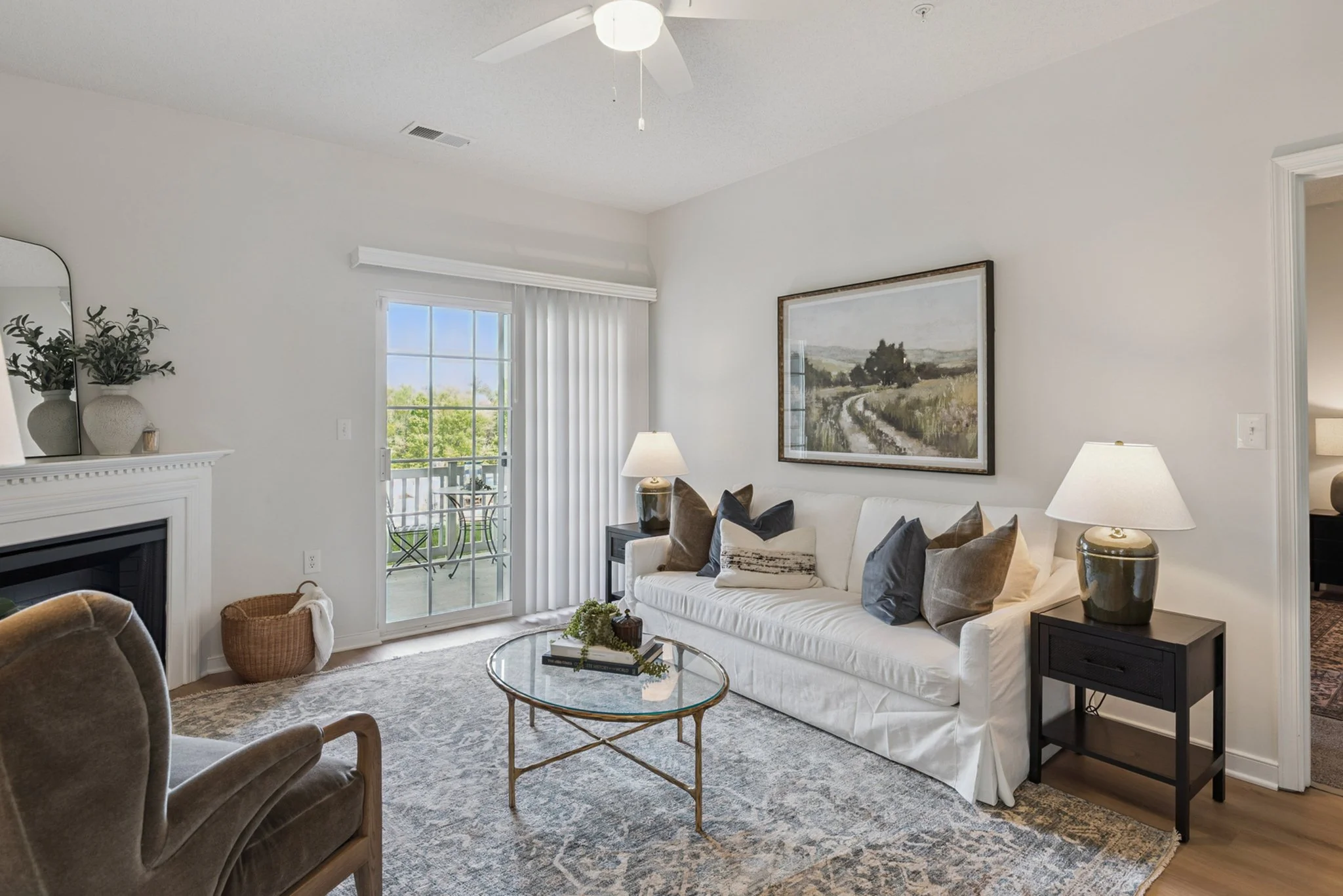 Living room with white sofa, decorative pillows, two side tables with lamps, a round glass coffee table, framed landscape painting, large window with outdoor view, vertical blinds, ceiling fan, fireplace with decor, armchair, and patterned area rug.