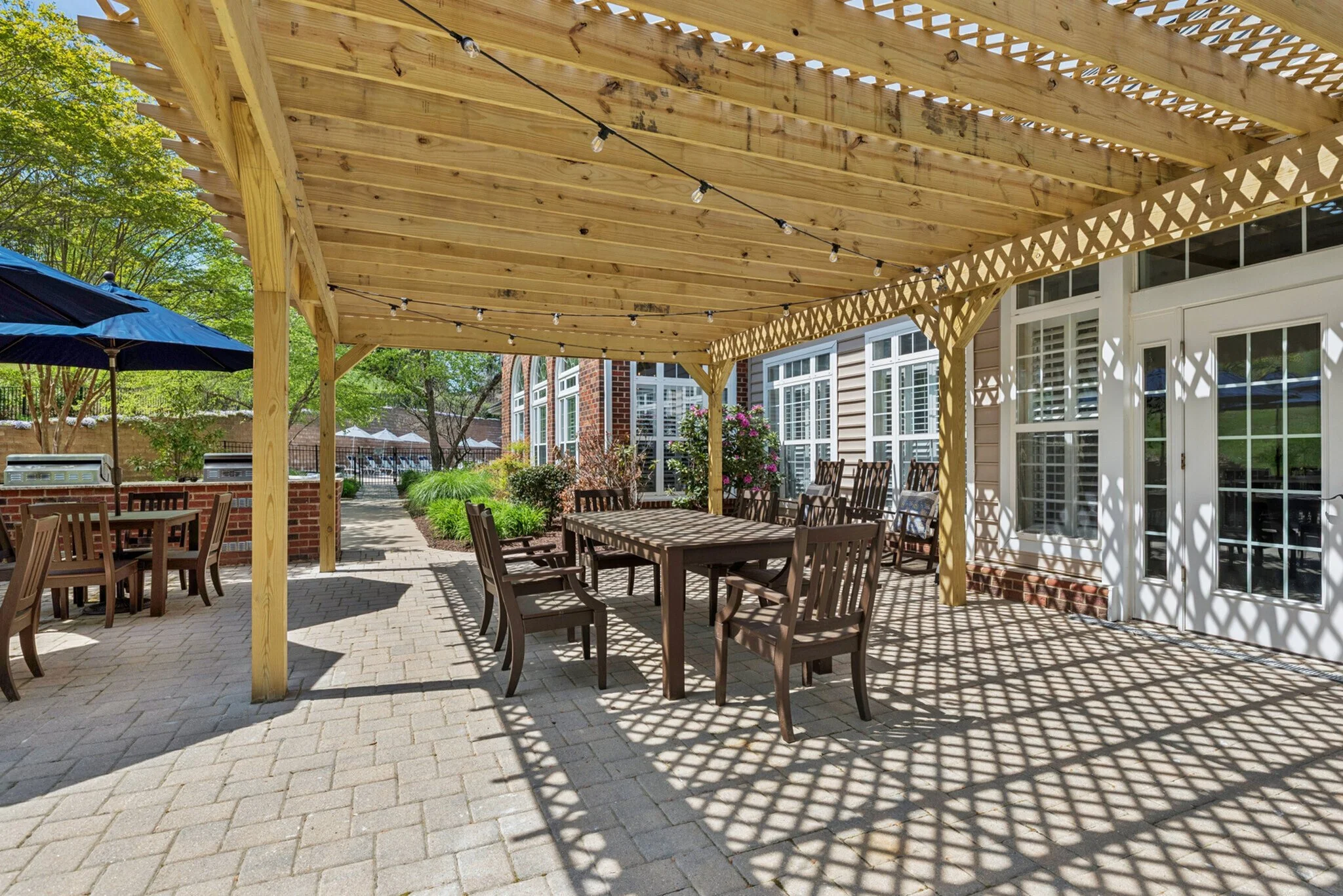 A backyard patio covered by a wooden pergola with string lights, surrounded by trees and a house with large windows and sliding doors.