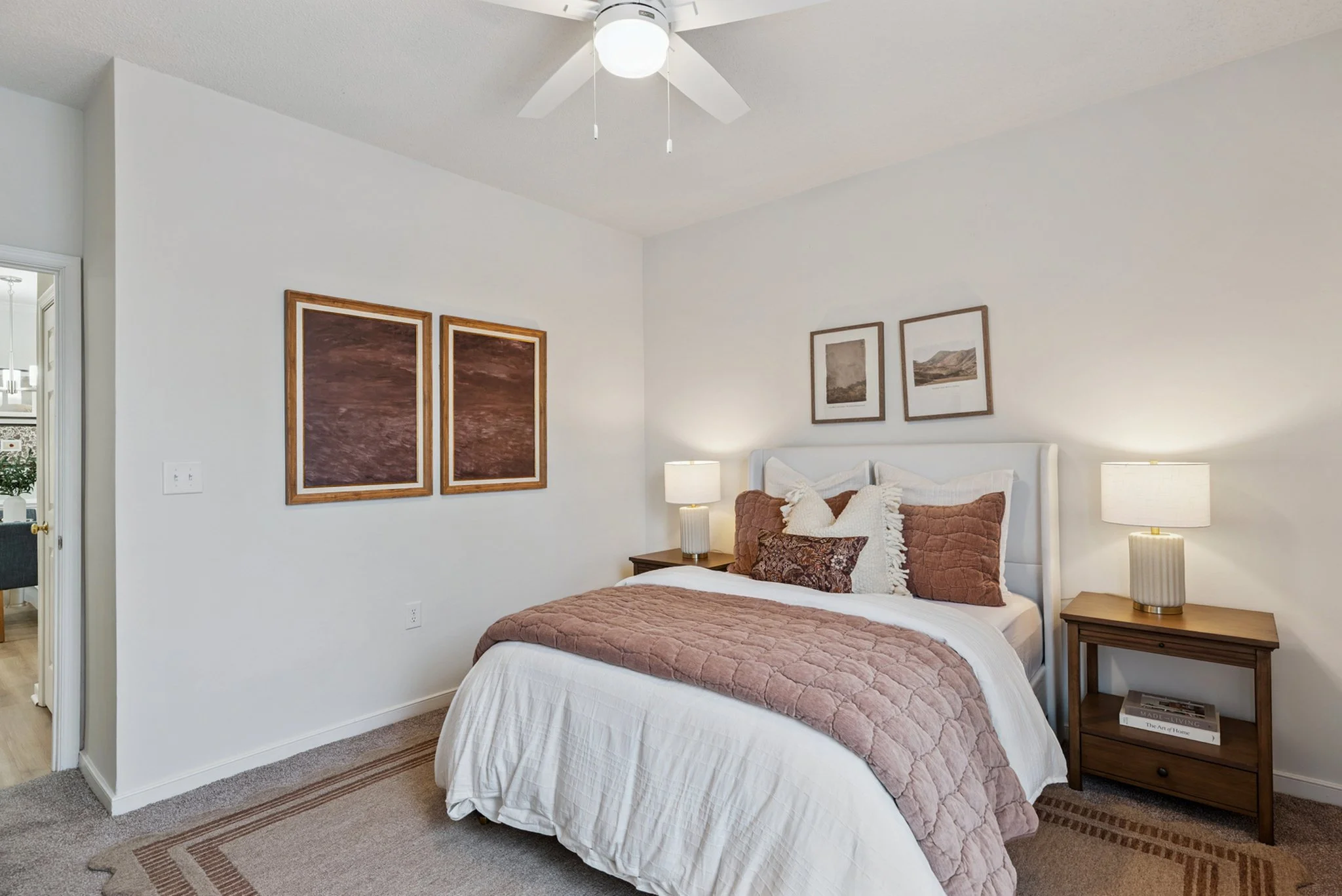 A bedroom with a white bed, brown pillows, a quilt, and framed artwork on the walls. Two bedside tables with lamps are on either side of the bed. There is a ceiling fan with light in the room.