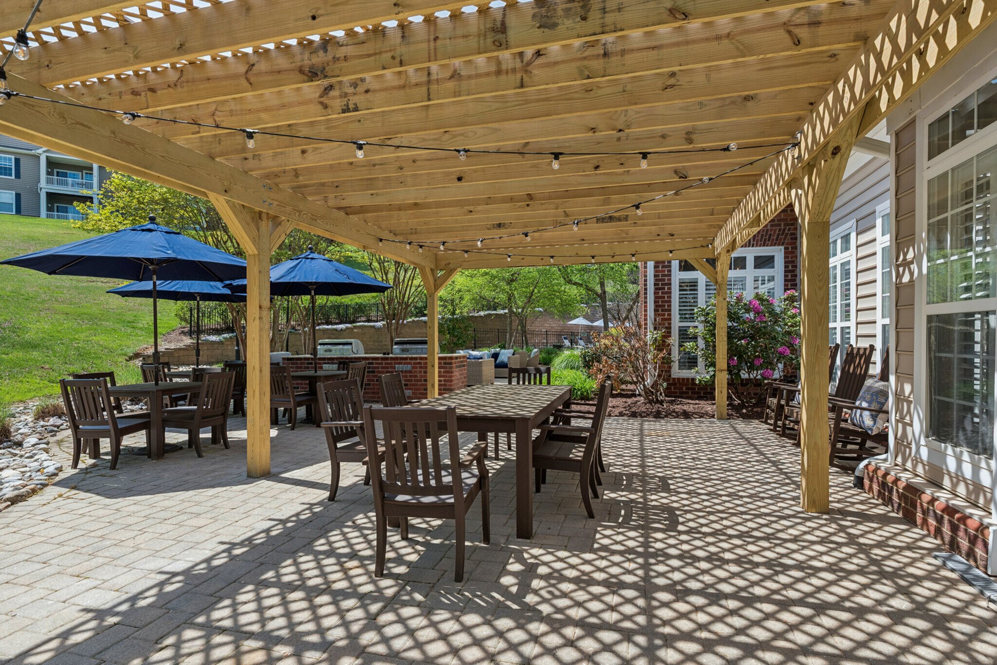 Backyard patio with wooden pergola overhead, string lights, outdoor dining tables and chairs, blue patio umbrellas, landscaped garden with flowering bushes and trees, brick wall, and neighboring houses in the background.