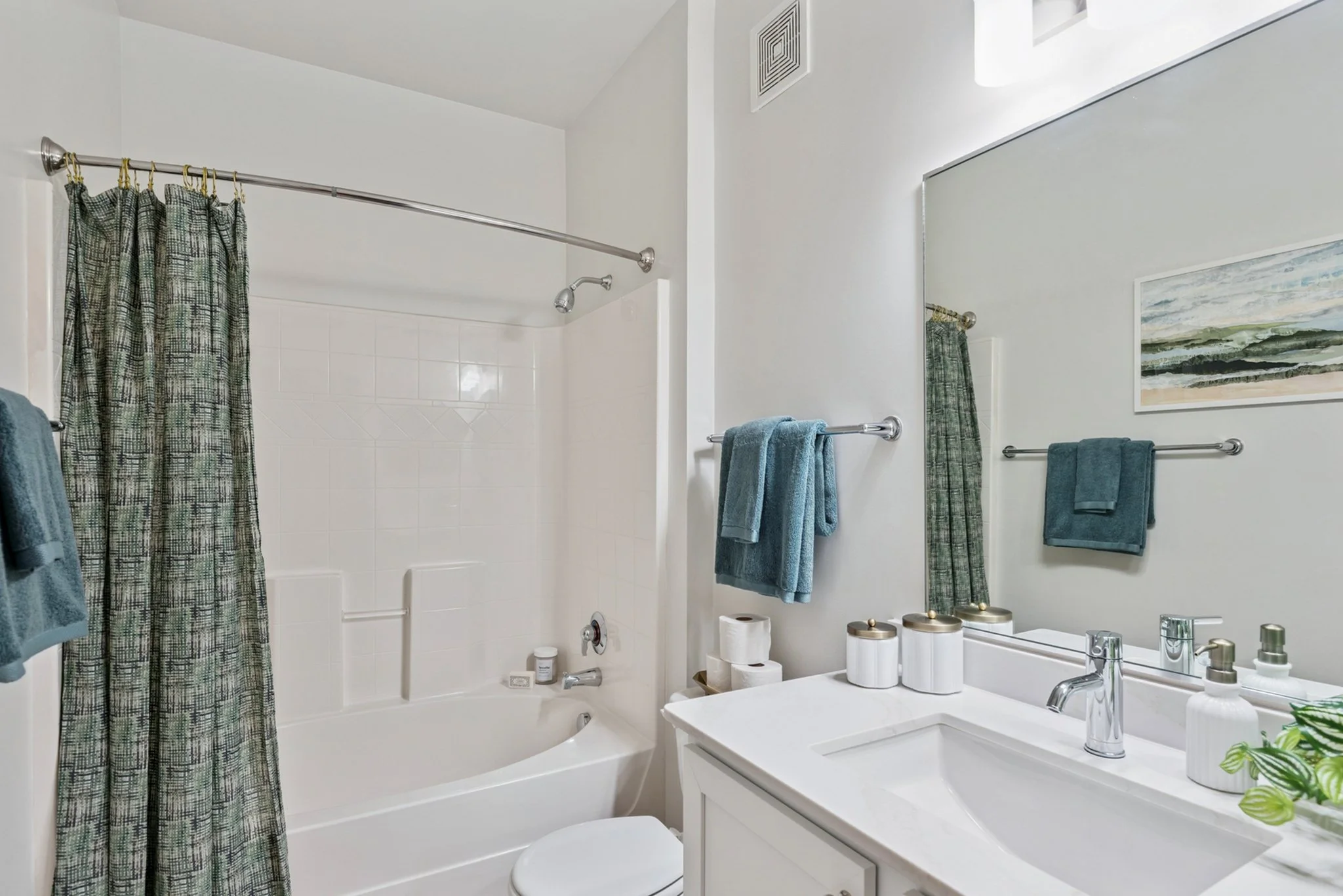 A clean white bathroom with a bathtub, shower area with a patterned green and black shower curtain, and a white vanity with a large mirror. There are teal towels hanging on a silver towel bar, and a framed watercolor landscape on the wall.