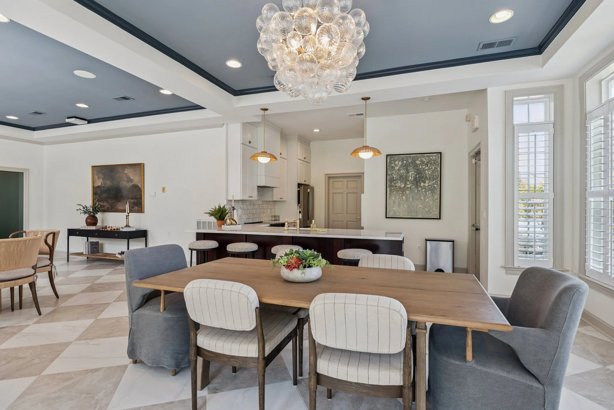 A modern open-concept living space featuring a dining area with a wooden table and various upholstered chairs, a kitchen with white cabinetry, a large chandelier made of glass globes, and windows with white shutters.