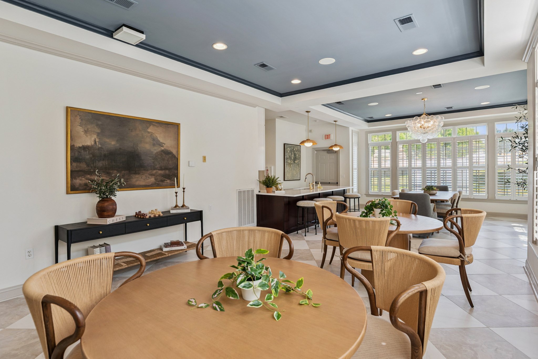 Bright dining area with round wooden table, surrounding chairs with woven backs, a potted plant in the center, large windows with white shutters, a chandelier, and a kitchen bar with stools.