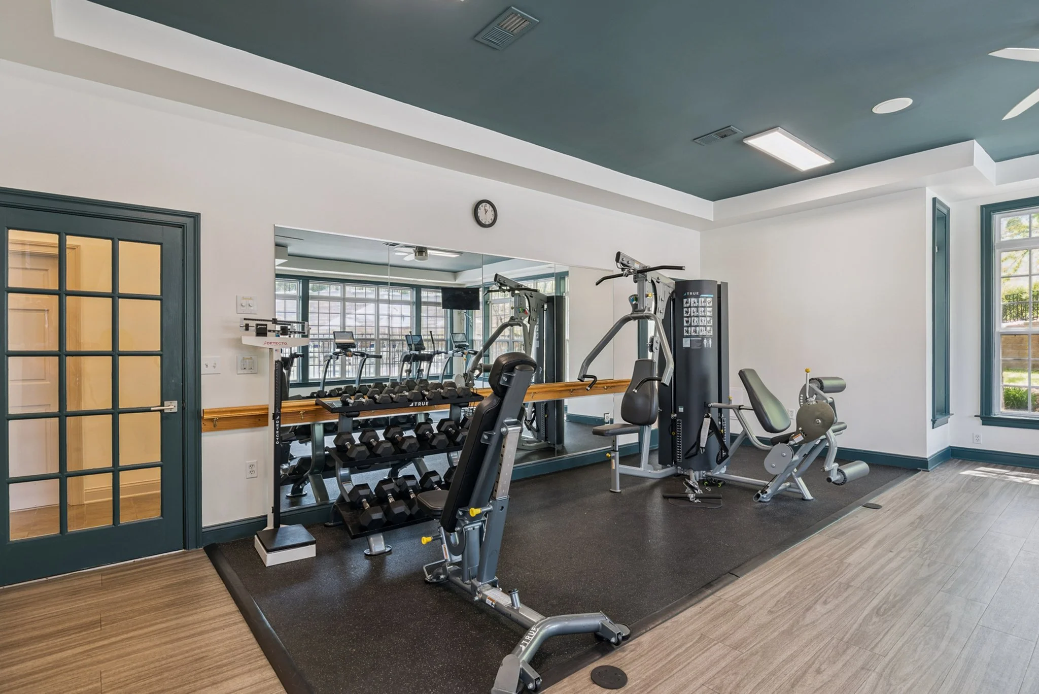 A fitness room with various exercise equipment including dumbbells, treadmills, and weight machines. There is a large mirror on the wall and windows letting in natural light.