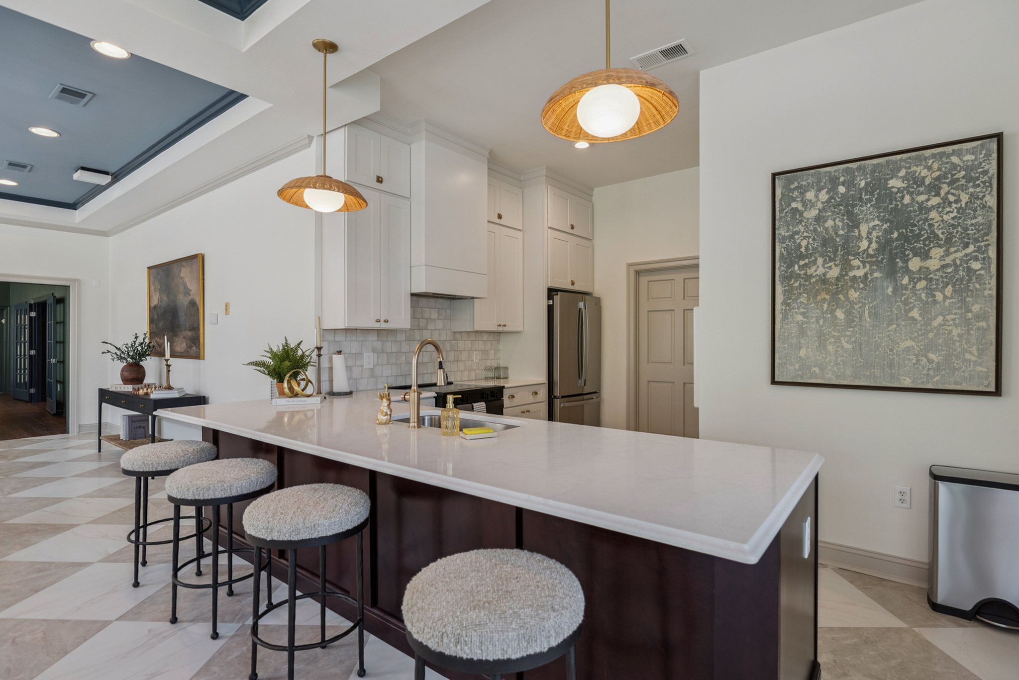 Modern kitchen with white cabinets, stainless steel refrigerator, marble island with four stools, pendant lights, and a large framed abstract artwork on the wall.