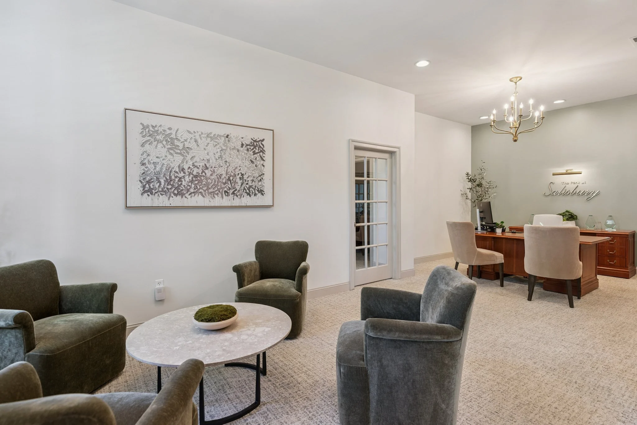 A cozy living room with a round coffee table surrounded by six plush armchairs, a white wall with a large framed abstract artwork, a window, and a desk with chairs near a wall with decorative lettering that says 'Salisbury'. A chandelier hangs from t