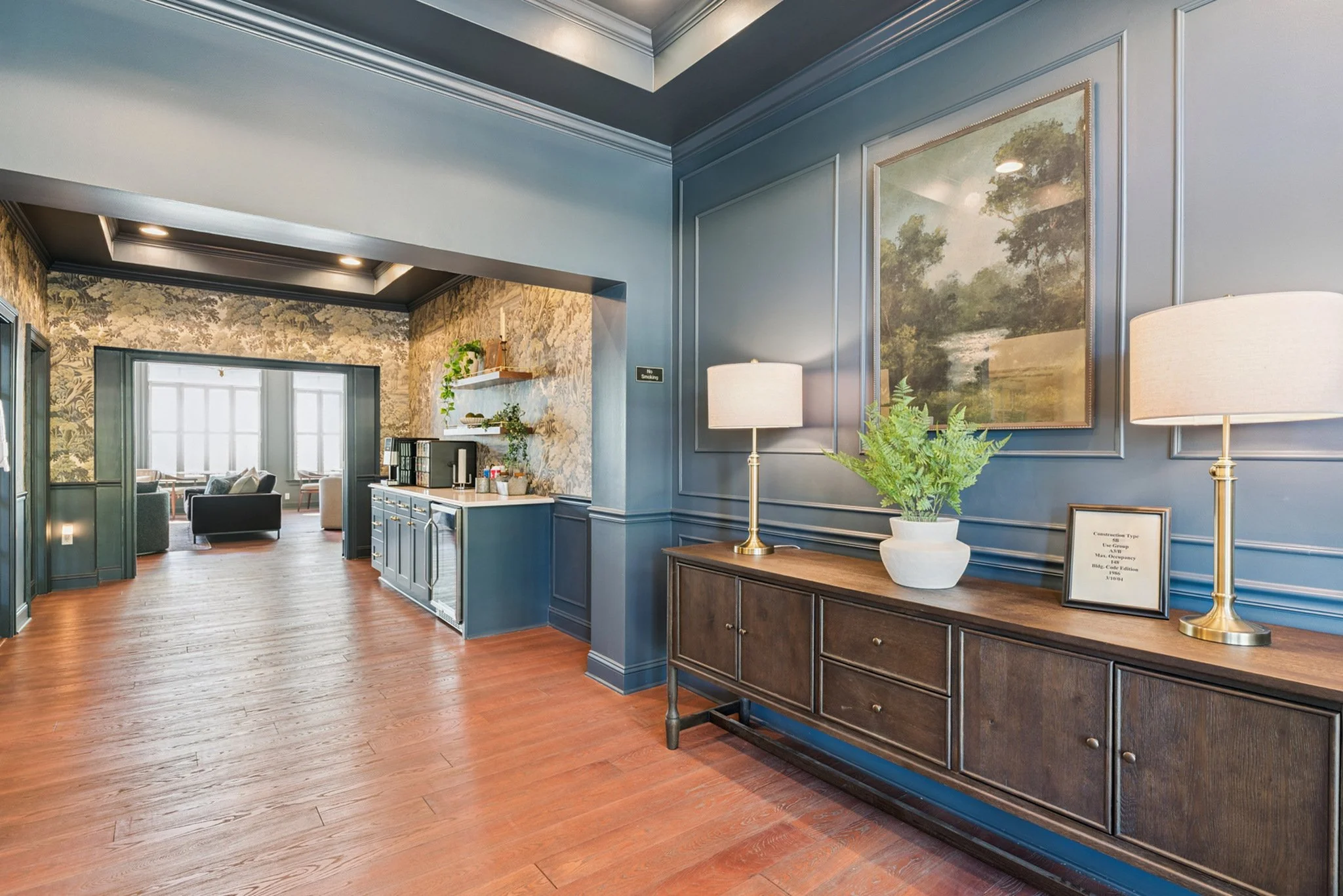 Interior of a room with blue paneled walls, framed landscape paintings, two table lamps with white shades, a dark wooden sideboard with plants and framed documents, and a view into a sitting area with sofas and large window.