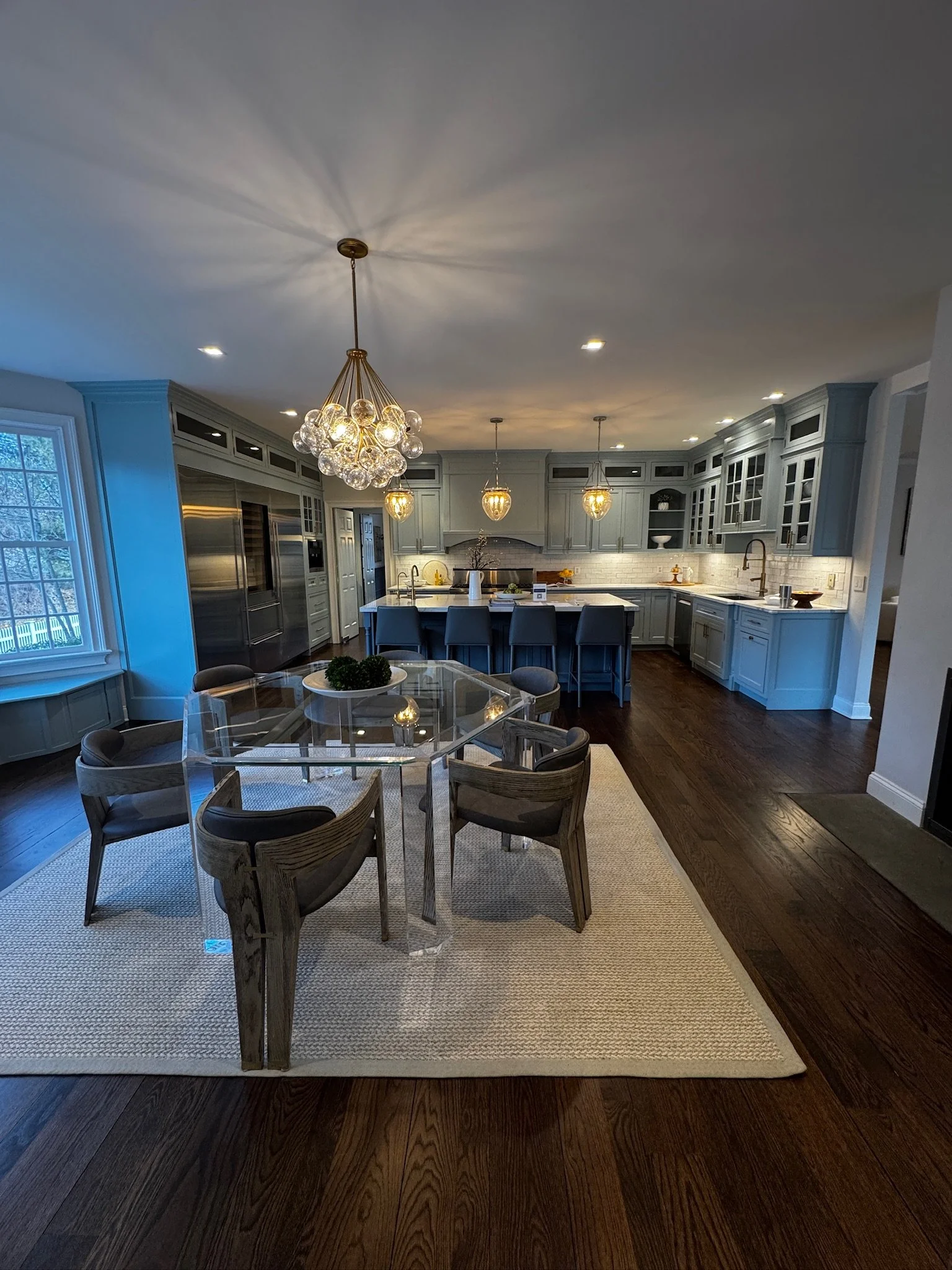 Open-concept kitchen and dining area with hardwood floors, modern cabinetry, and pendant lighting, featuring a round glass dining table with six chairs and an island with bar stools.