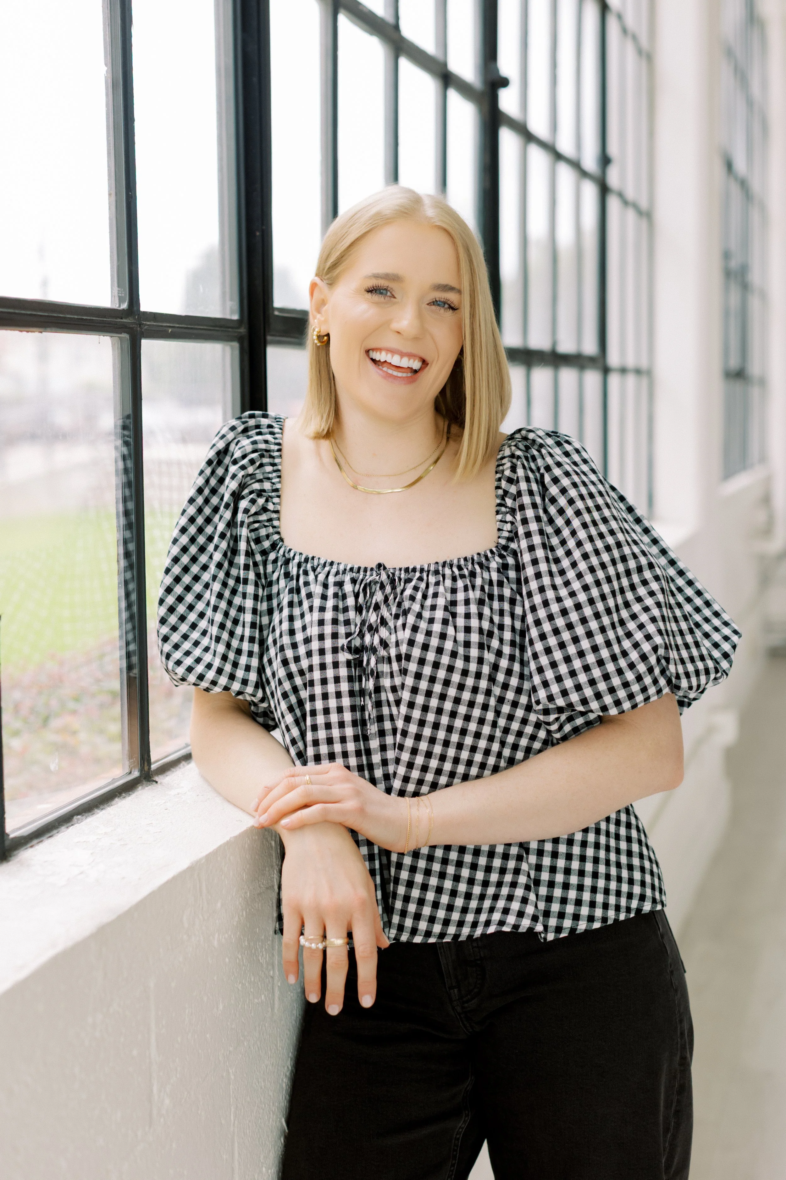 A young woman with blonde hair, wearing a black and white checkered top and black pants, smiling while leaning on a windowsill in a bright, modern space. EMDR Anxiety Counseling LPC Houston Heights Dallas Texas