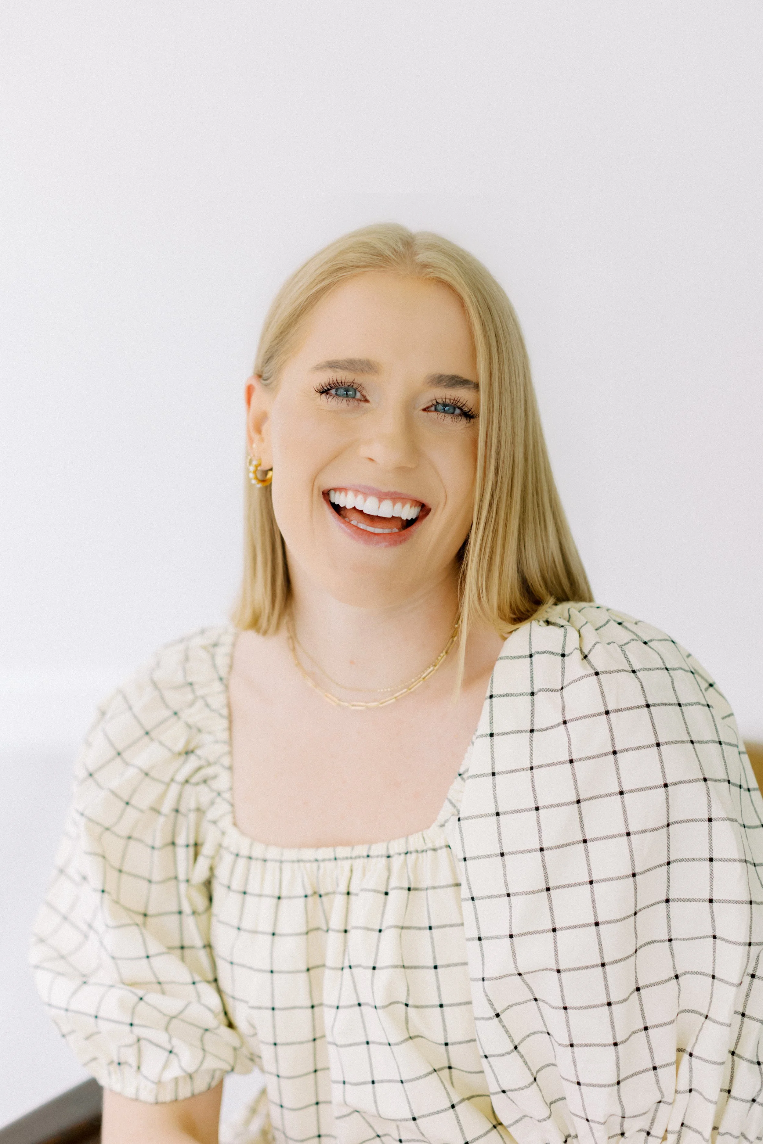 A young woman with blonde hair smiling and wearing a cream-colored dress with black checkered pattern, gold earrings, and layered necklaces, against a plain white background. Alive and Whole Wellness Houston Texas. Heights LPC.