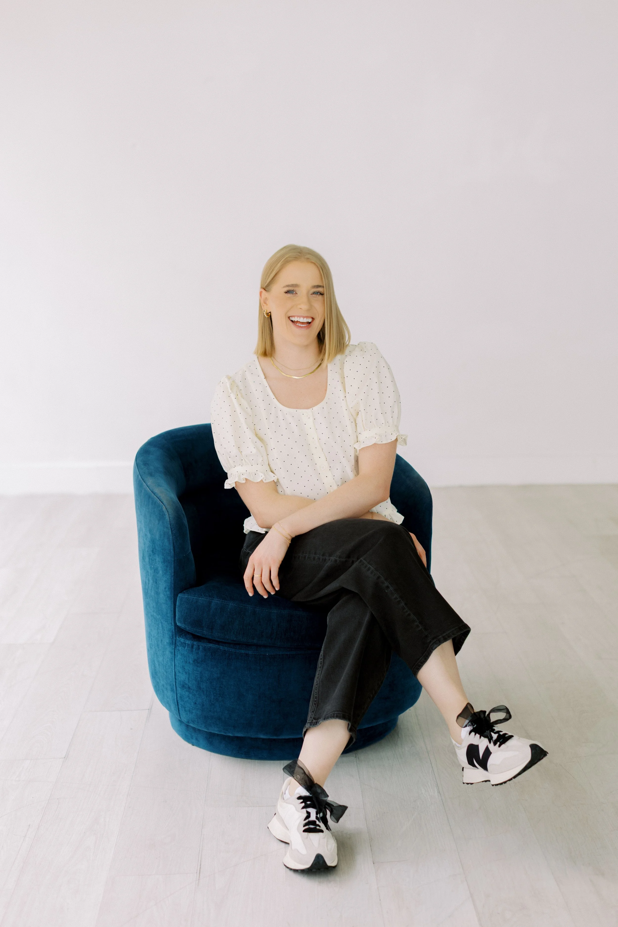A young woman with blonde hair, wearing a white polka dot blouse, black pants, and white sneakers with black laces, sitting on a blue velvet chair in a minimalistic room with light-colored hardwood floors and white walls, smiling and looking at the c