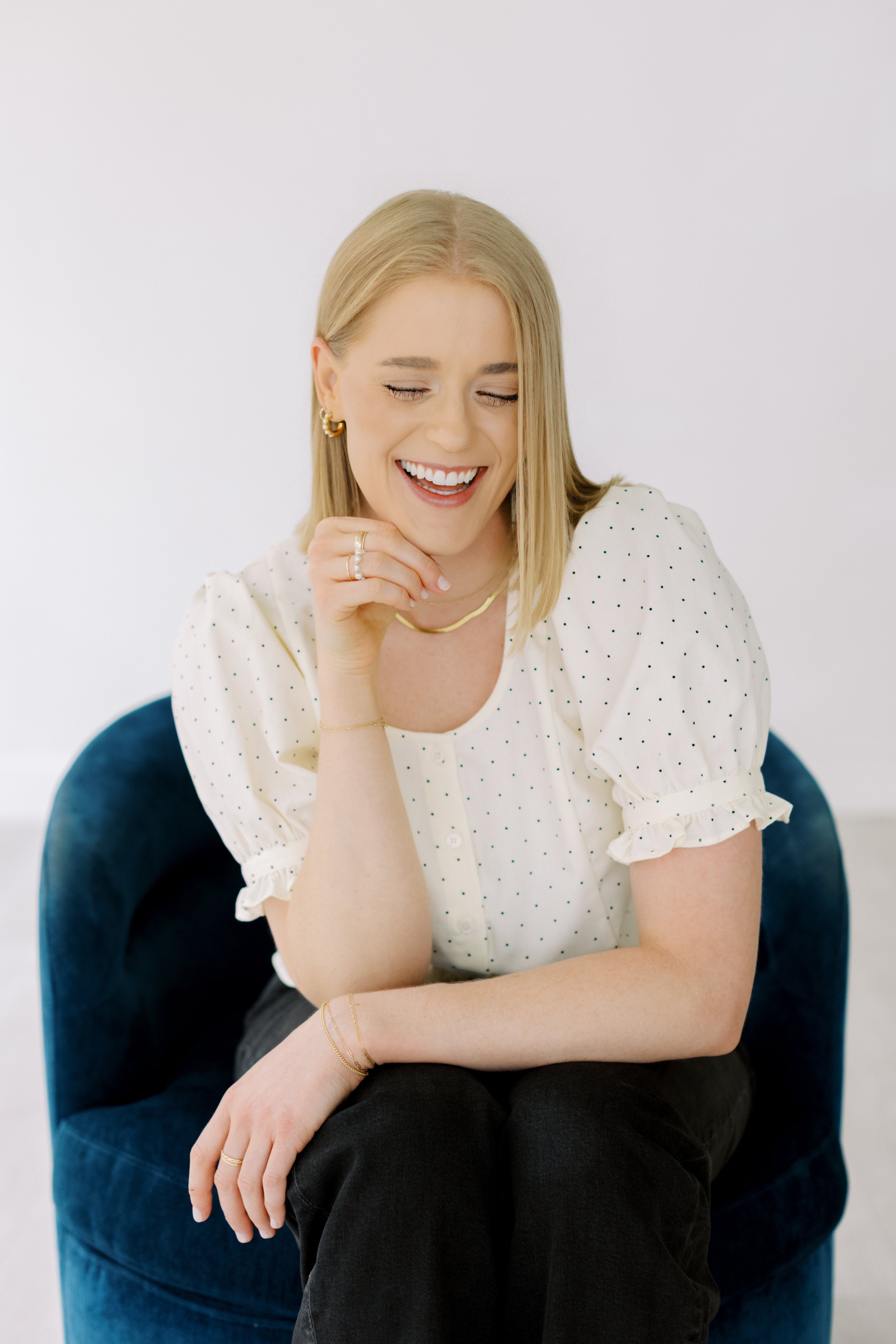A woman with blonde hair sitting in a blue velvet chair, smiling with eyes closed, wearing a white blouse with black polka dots, gold jewelry, and black pants. Christian Counseling. Houston Texas. Alive, whole, and free. Heights. Emily Rucker LPC. 