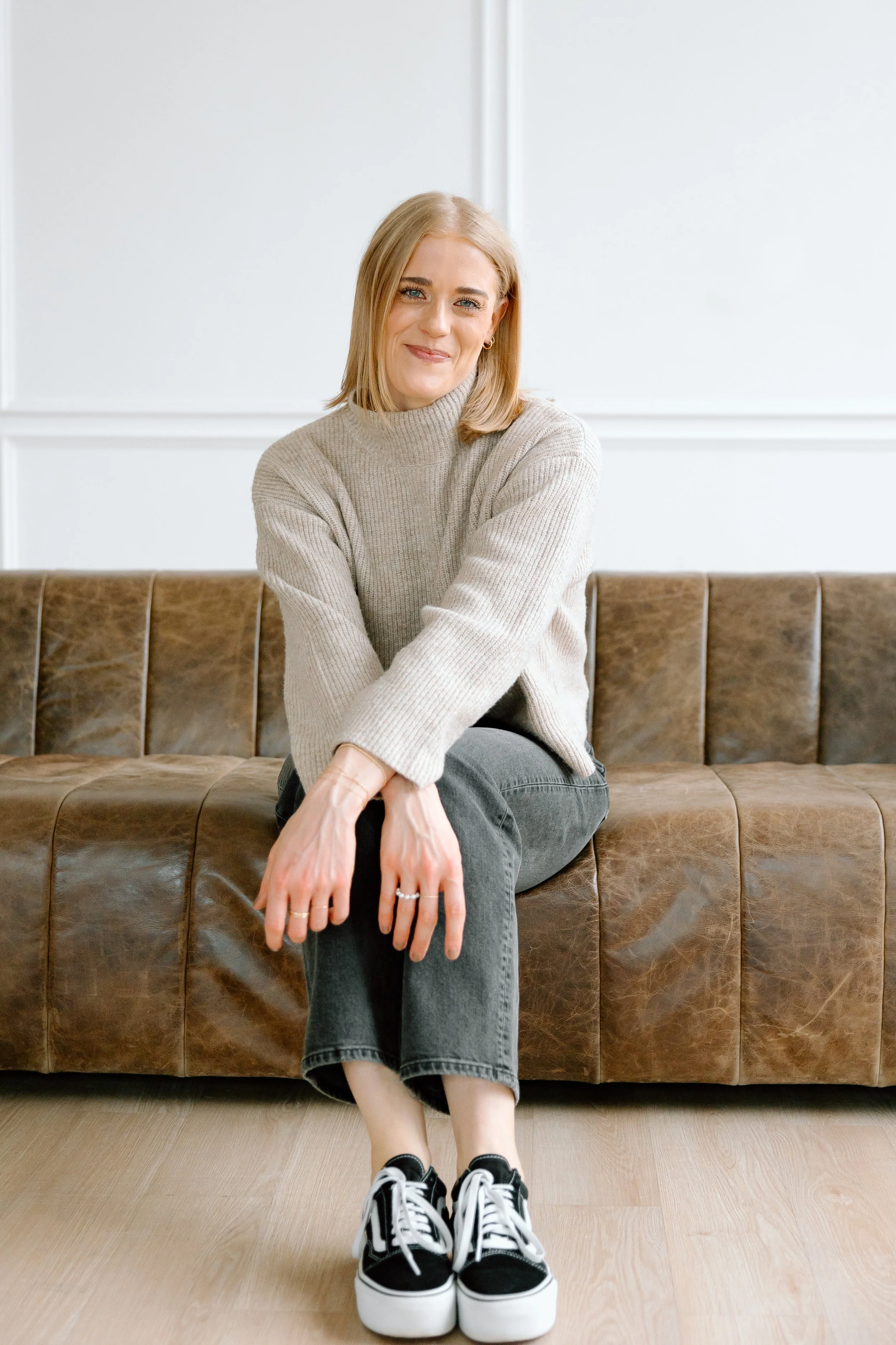 Woman sitting in a relaxed pose on a brown leather couch, wearing a beige sweater, black jeans, and black sneakers.
