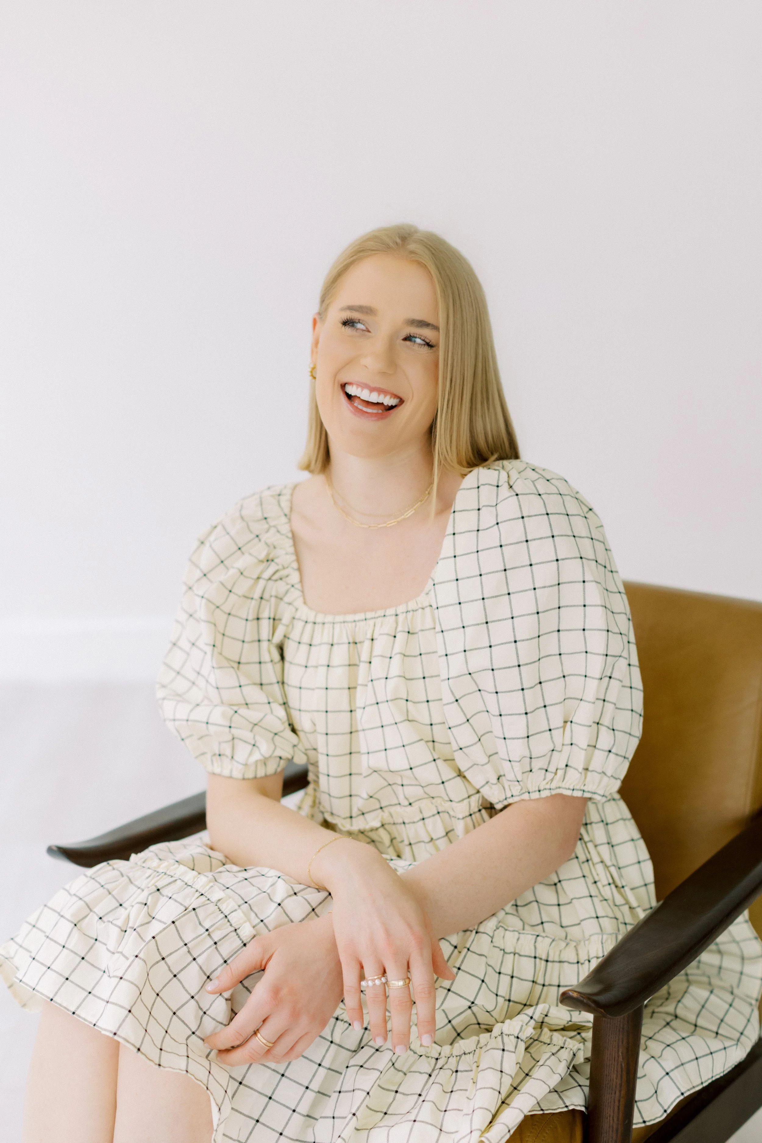 A young woman with blonde hair, wearing a beige dress with a black grid pattern, sitting on a wooden chair, smiling and looking to her left. Emily Rucker M.A. LPC. Houston Texas. Alive and Whole Wellness. Christian Counseling. EMDR. Trauma. Anxiety.