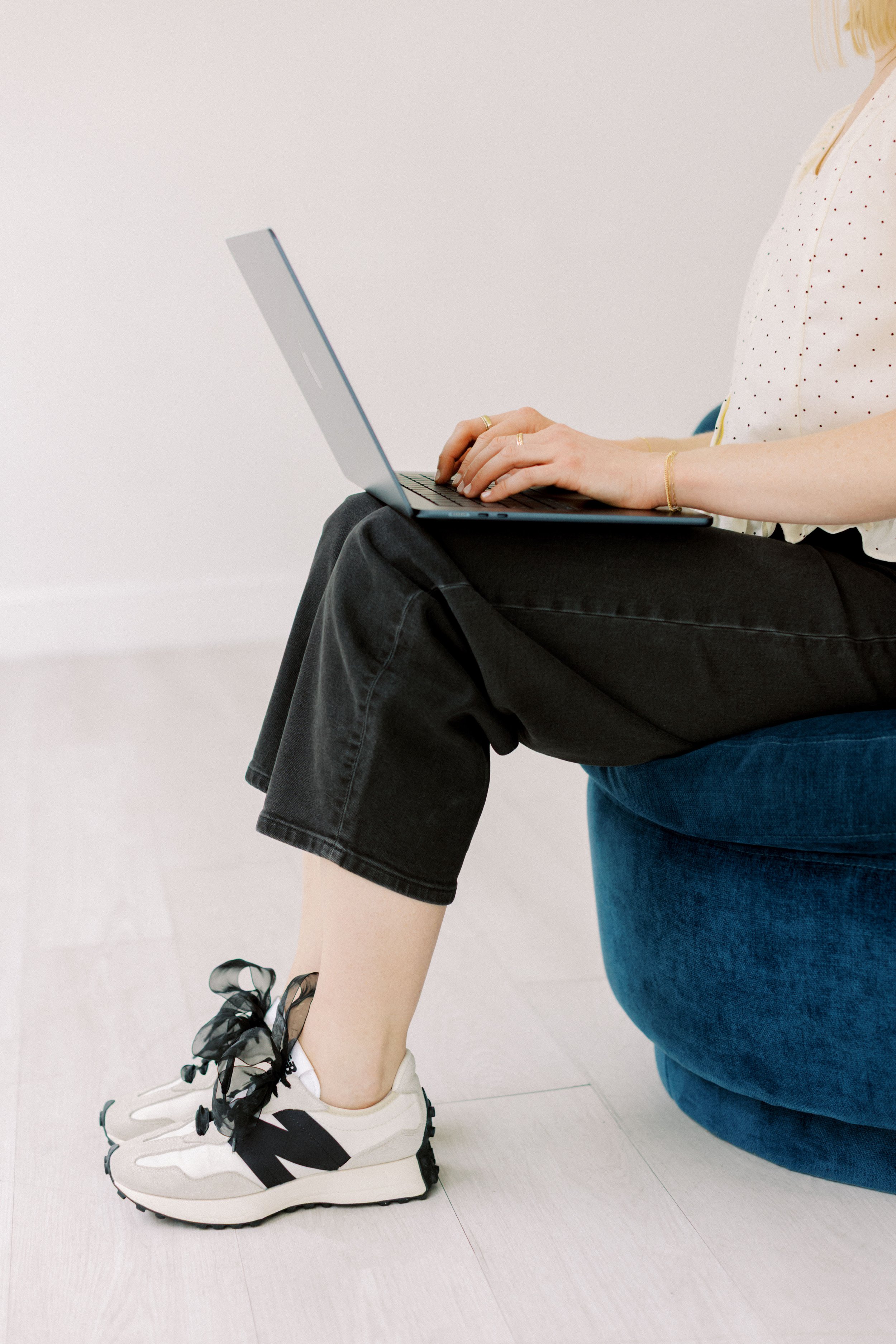 Person sitting on a person's lap, using a laptop, wearing white sneakers with black accents, black shorts, a beige polka-dot shirt, and a blue skirt, with a plain white background. Emily Rucker M.A. LPC. Houston Texas.