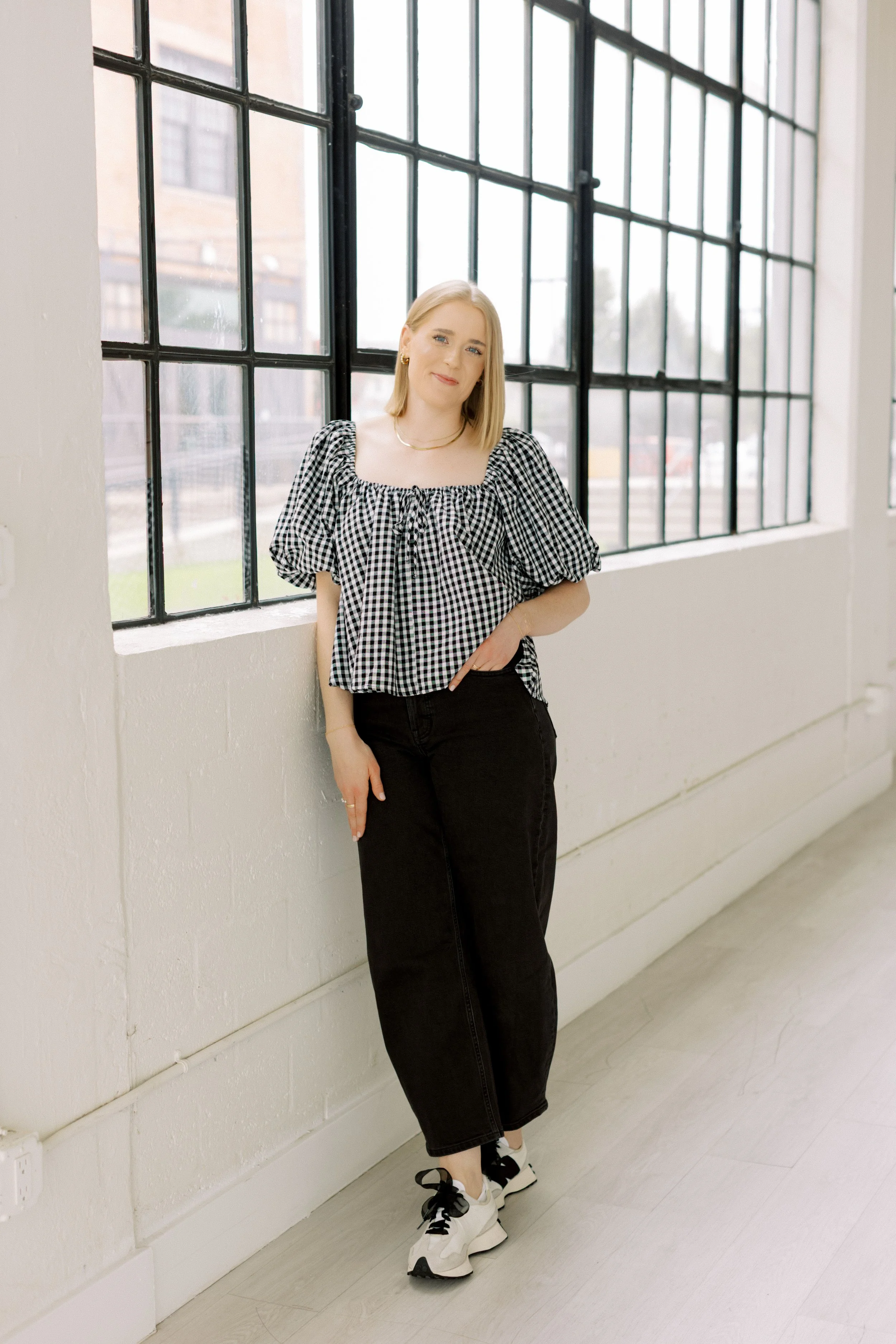 A woman with blond hair in a straight style, wearing a black-and-white checkered blouse with puffed sleeves and a tie at the neckline, black high-waisted jeans, and white sneakers with black laces, standing indoors by a large window with black grid panes, and smiling softly.