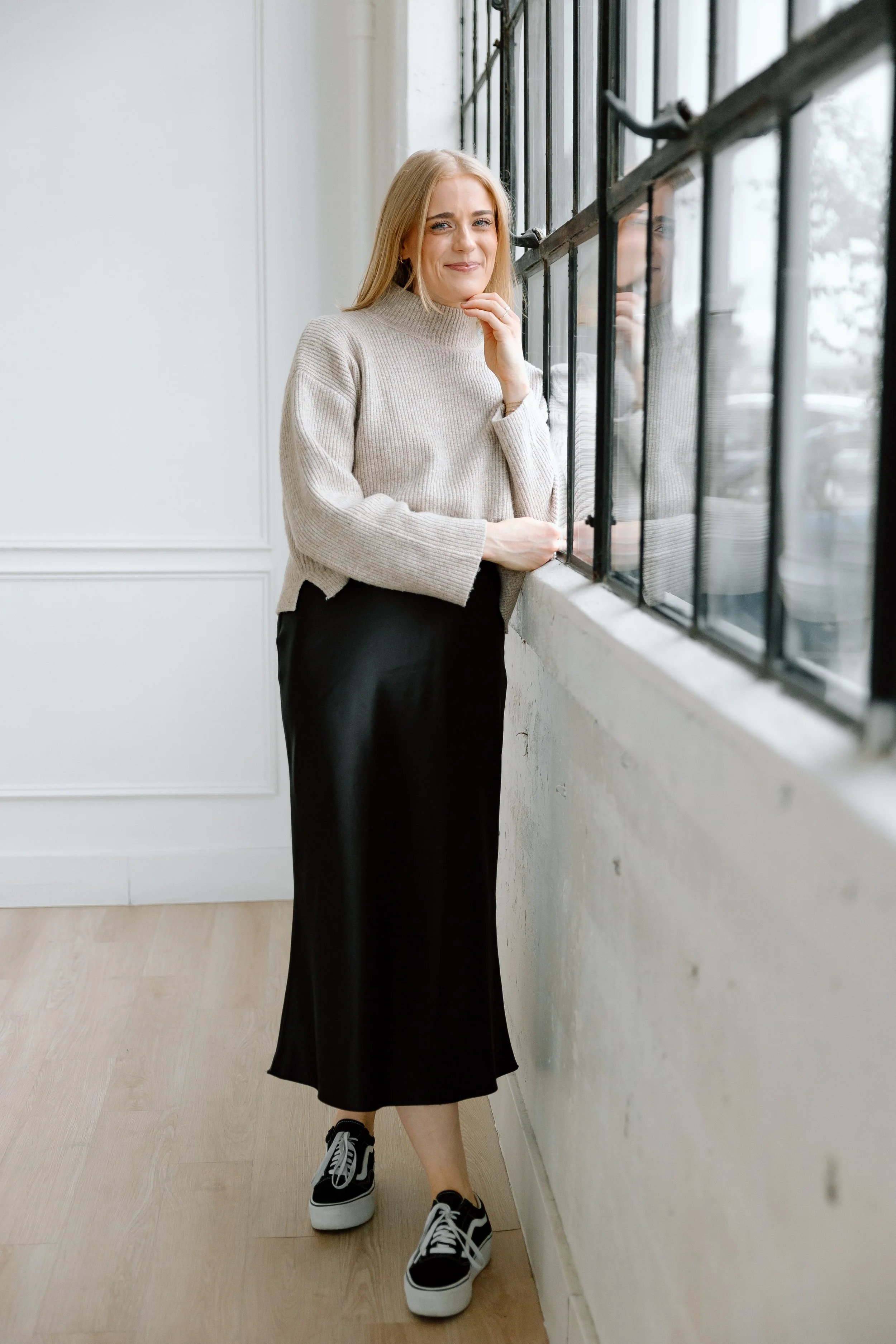Woman in beige sweater and black skirt standing by a window, smiling.