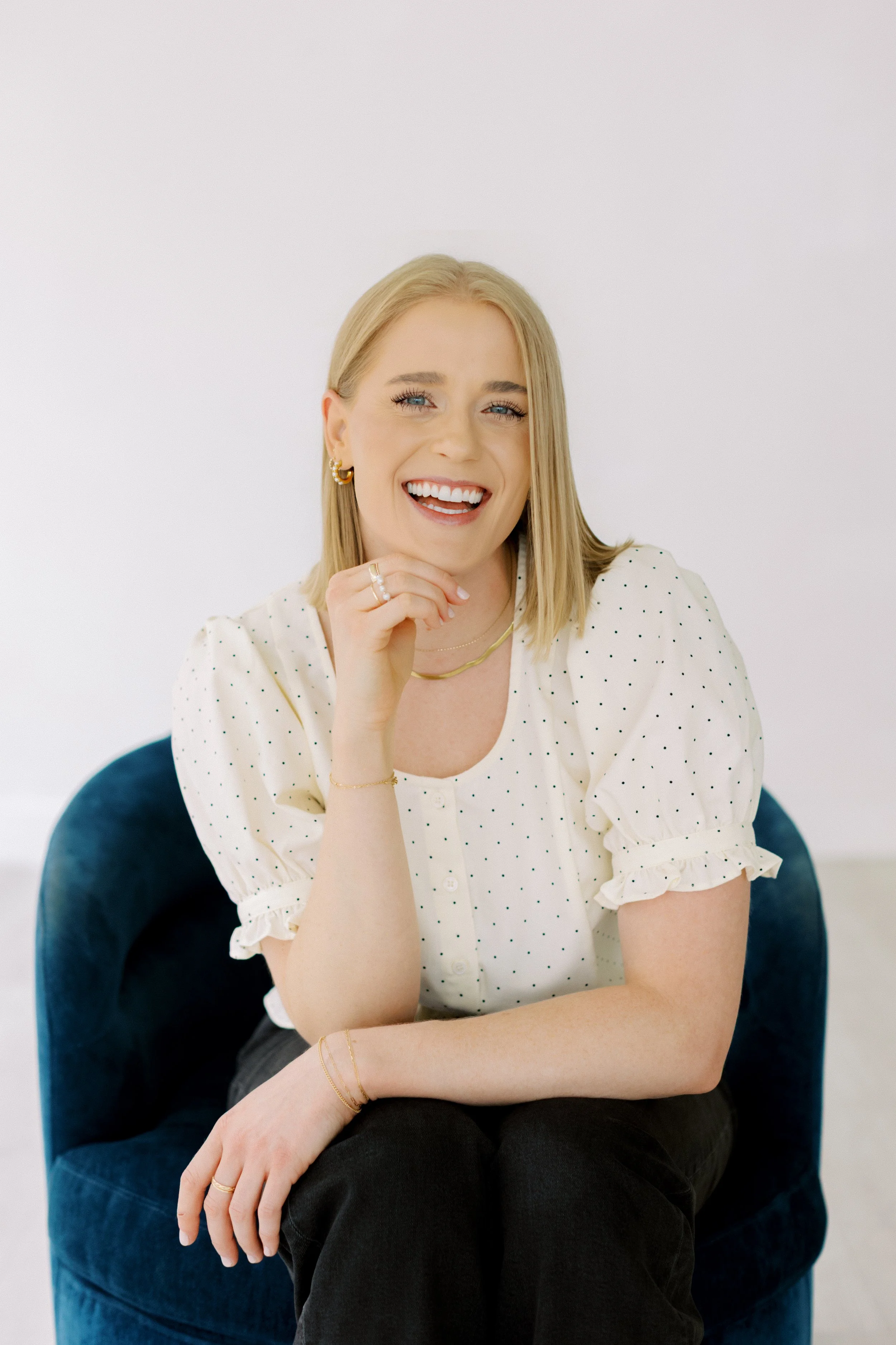 A smiling blonde woman with blue eyes, wearing a white polka dot blouse and gold jewelry, sitting on a blue velvet chair against a plain white background. Christian Counseling. Houston Texas. Emily Rucker M.A. LPC. Alive and Whole Wellness. 